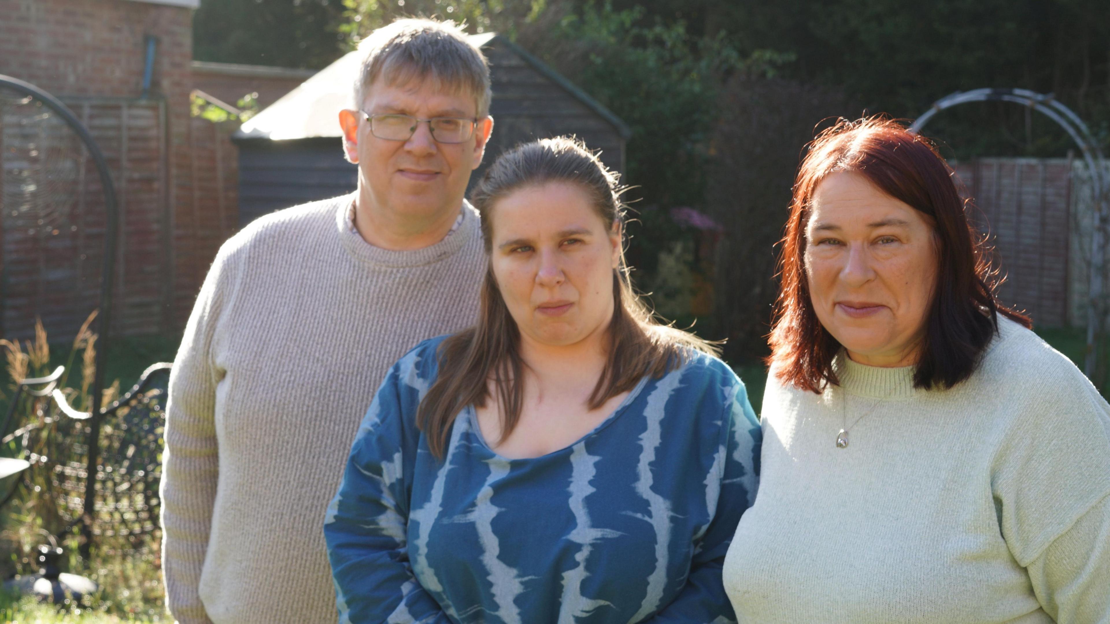 Adrian, Dawn and Judith are standing next to each other in the garden. They are looking at the camera with neutral expressions. Adrian and Judith are standing either side, wearing light-coloured jumpers and Dawn is in the middle with a blue patterned dress.