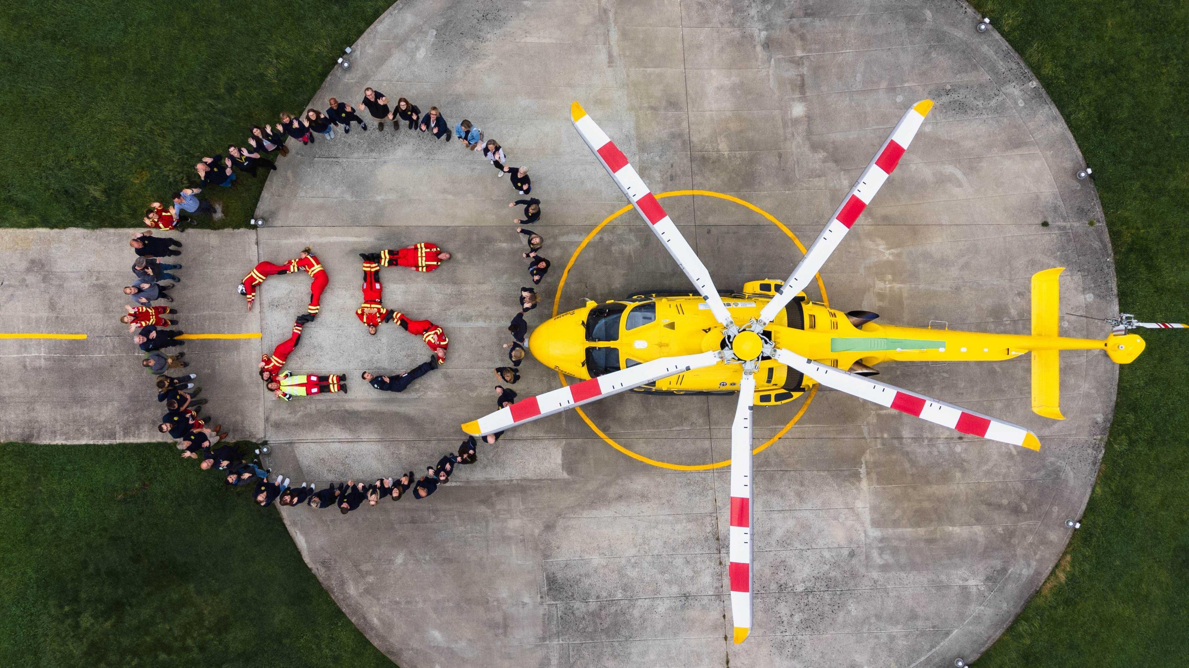 Aerial view of people standing in a circle around eight other people making the figure 25, next to a yellow helicopter.