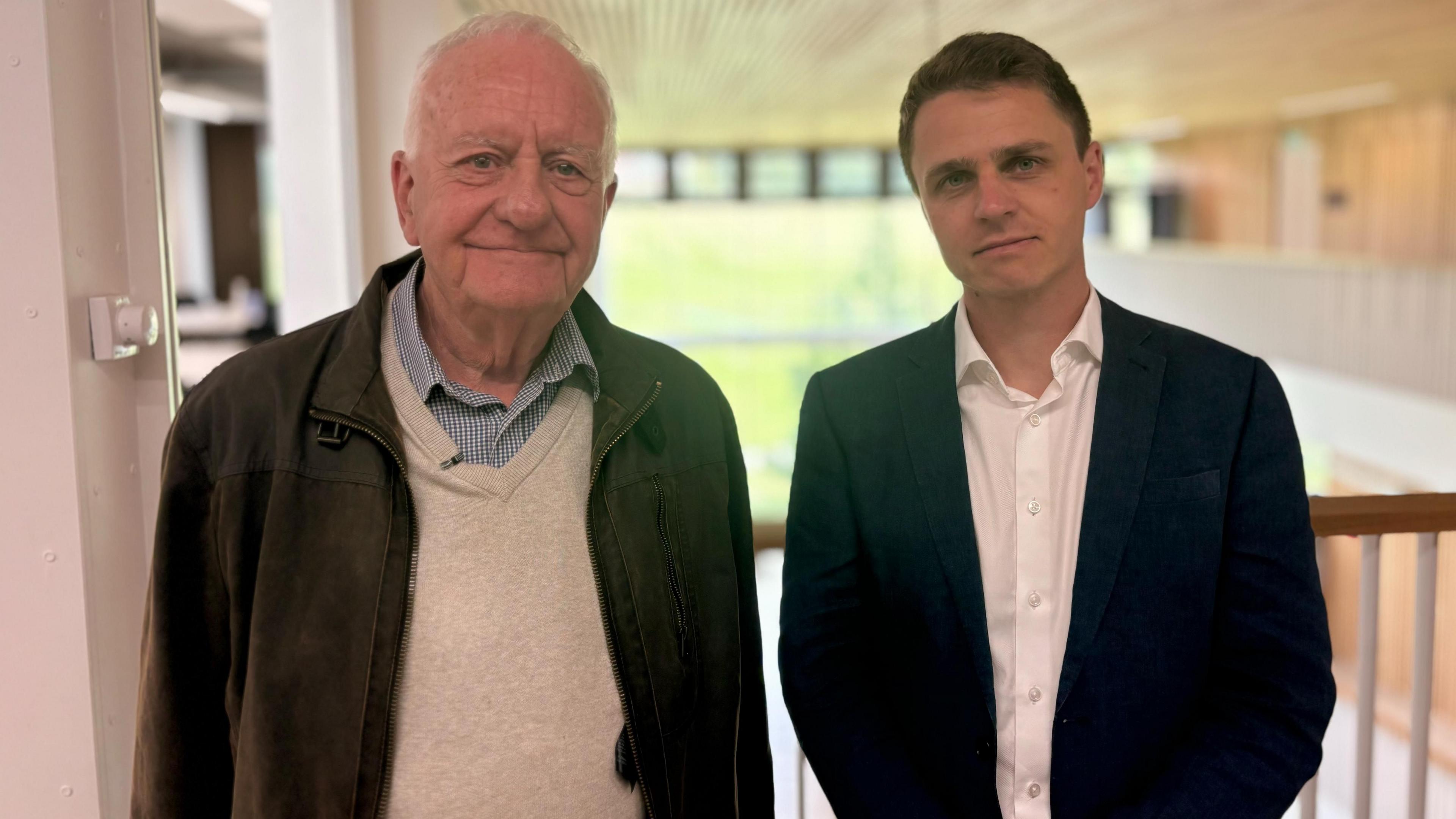 Steve Hindley (left) and Dr Michael Lambert (right) stand side by side on a mezzanine walkway indoors, with Steve wearing a brown leather jacket over a light jumper and check shirt, and Michael dressed in a dark suit jacket and white shirt. There is a softly blurred background of windows and interior walls.
