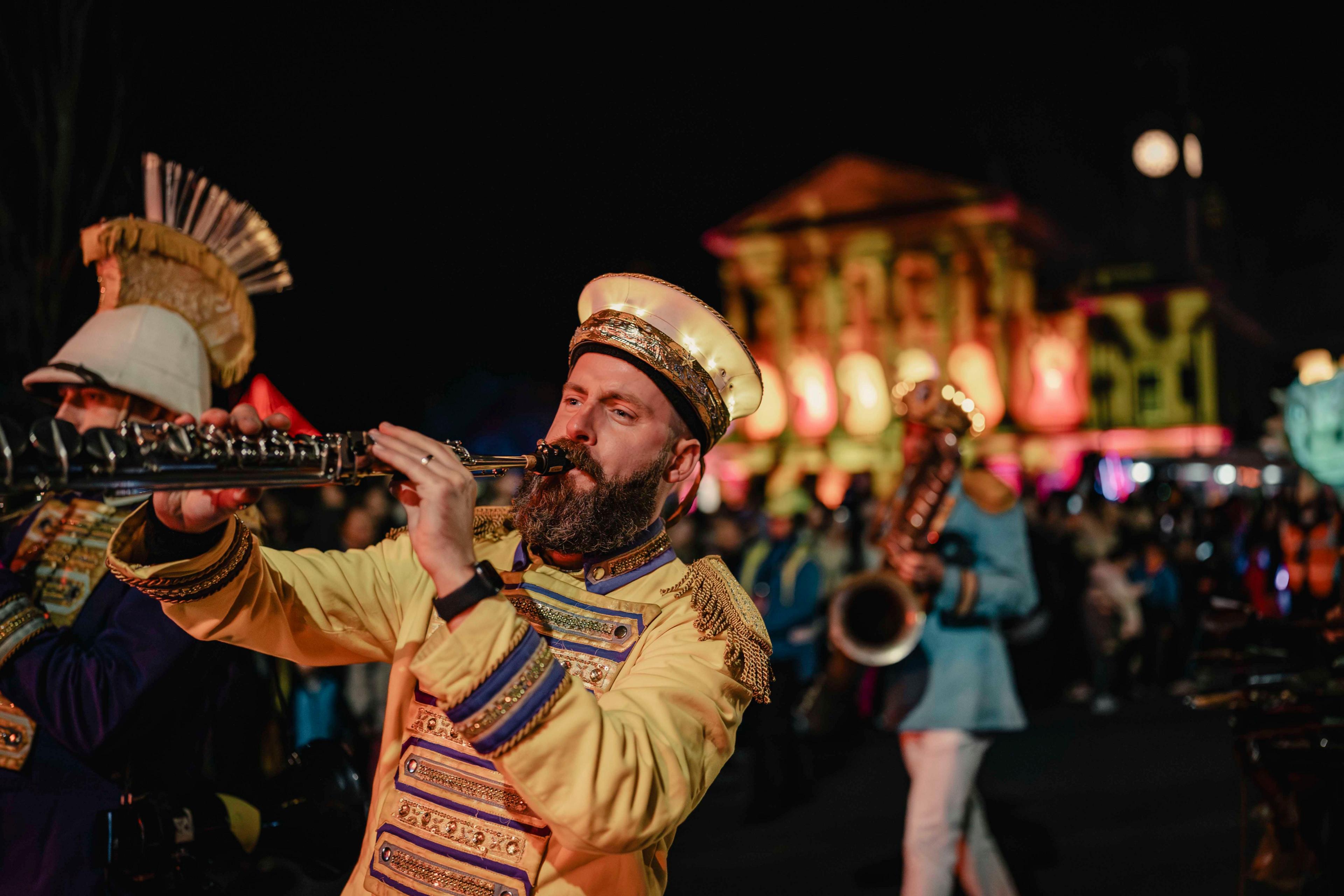 A number of woodwind players in yellow and blue regalia perform in a street parade. They are watched by crowds who are blurred in the background of the shot.