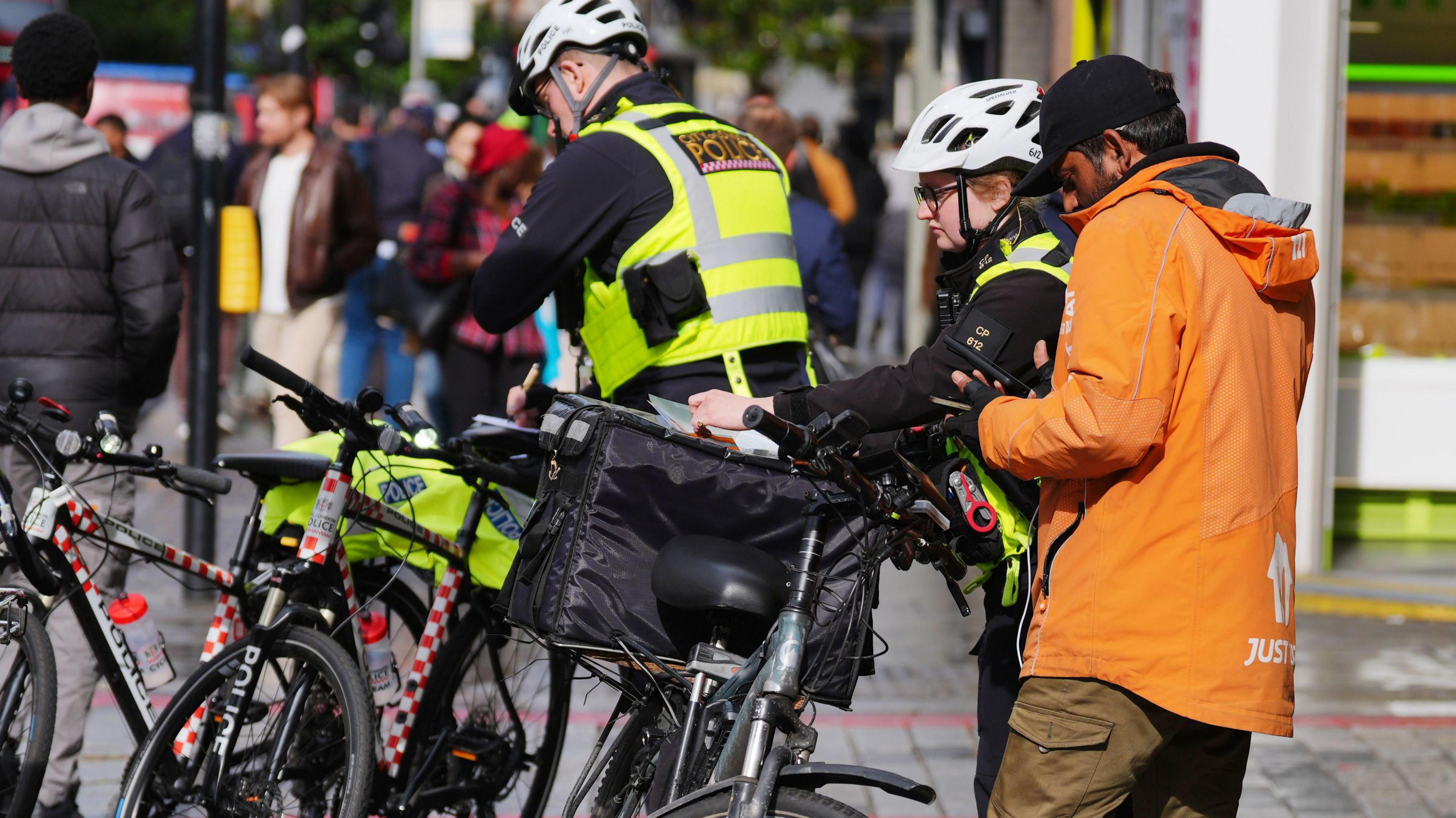 Two specialist cycling police officers stop a deliver rider wearing a brightly coloured Just Eat jacket. They appear to be examining the bike he was riding. Both police officers have high-vis jackets on and are wearing white helmets. 