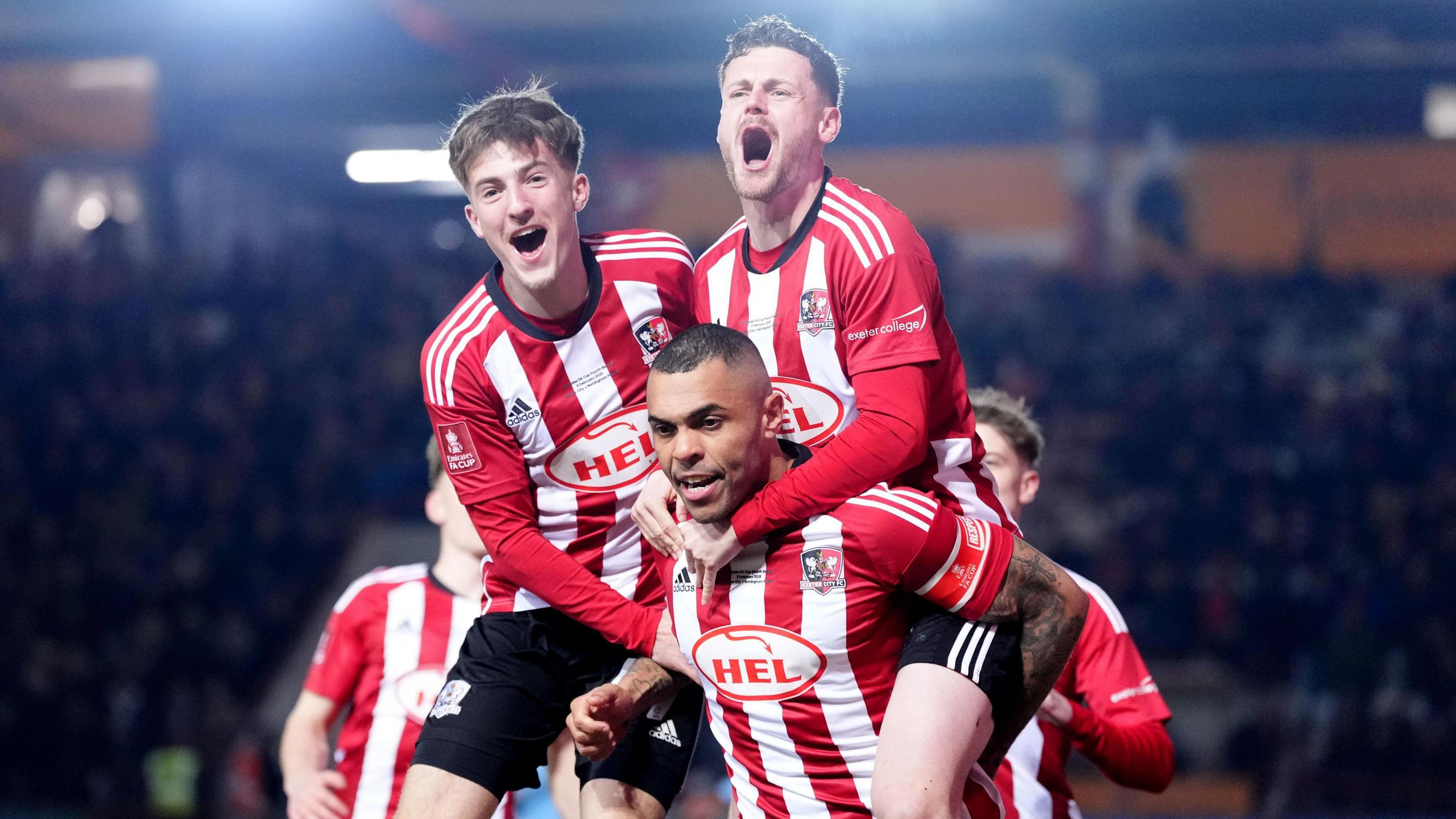 Exeter City's players celebrate scoring their side's first goal of the game during the Emirates FA Cup fourth round match at St James Park, Exeter. Three players with two more just visble in the background are wearing red and white striped long sleeve football jerseys. Two of the men appear to be jumping or on the back of the third. They both have their mouths open as if they are happily shouting or cheering. 