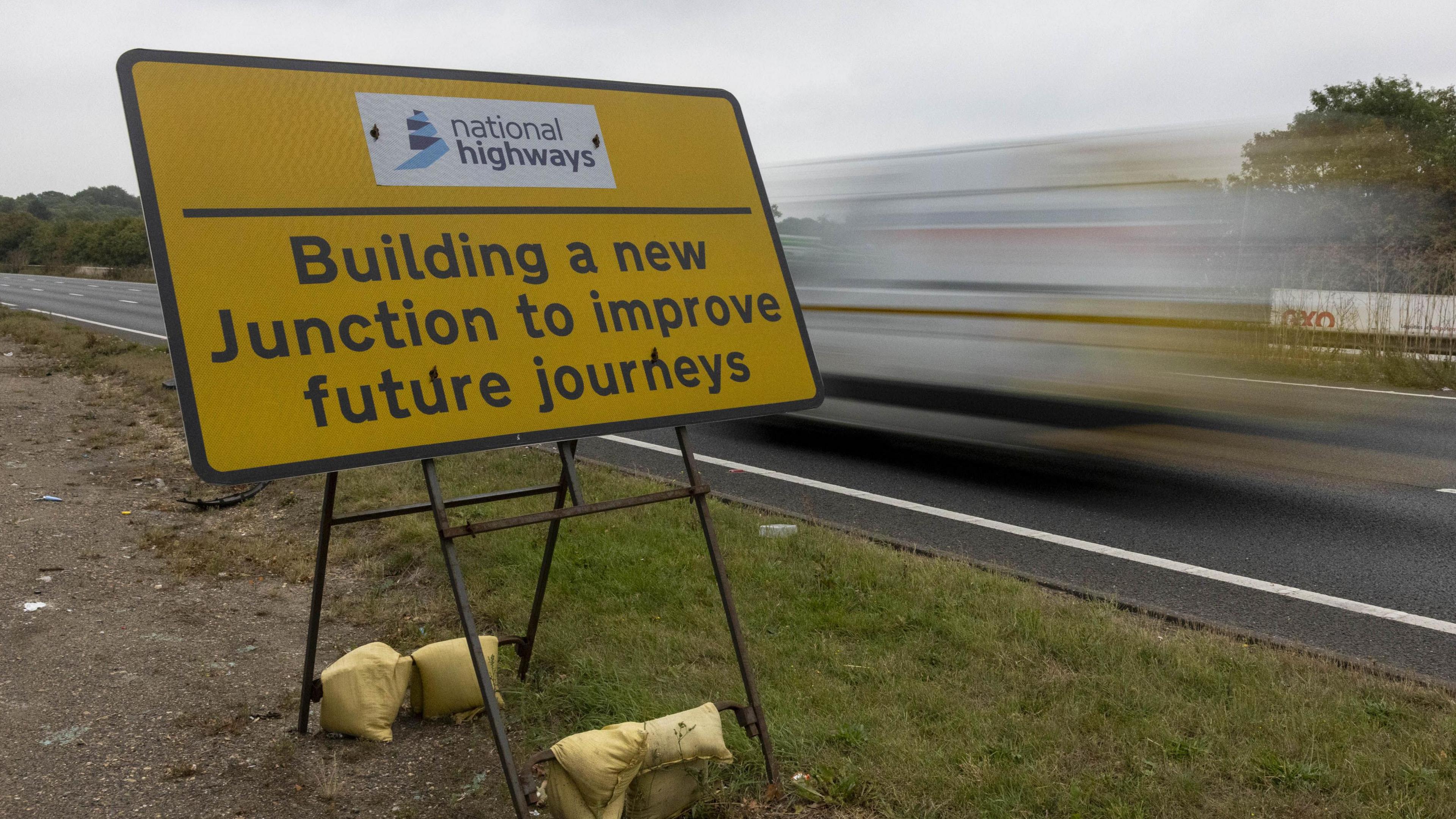 A yellow sign beside a road with the National Highways logo at the top, with the words 'building a new junction to improve future journeys' below. There is a blurred vehicle on the road to the reside of the sign