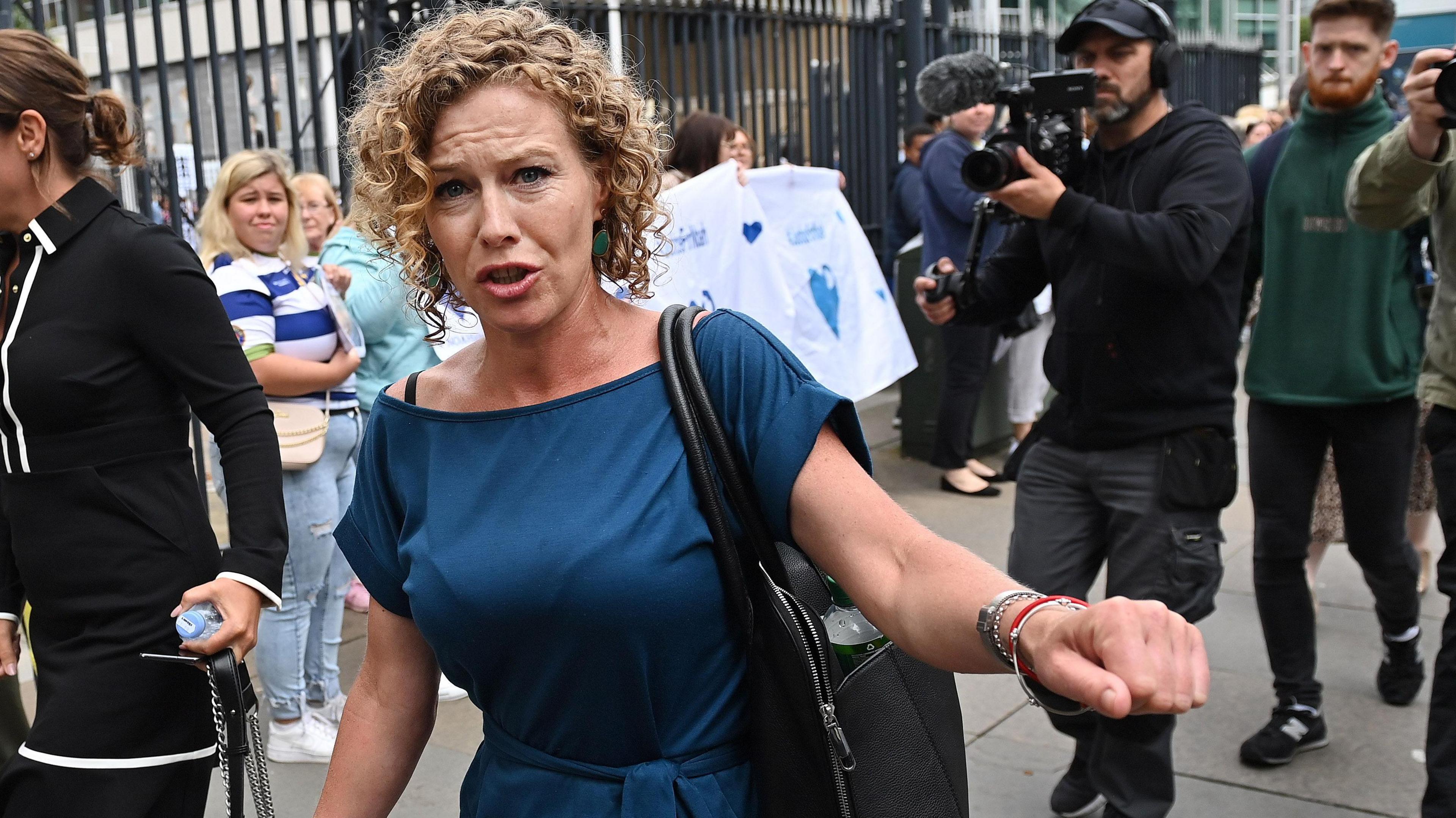 Fiona is outside court. She looks upset. She's wearing a blue dress and has a black bag. She's surrounded by people, including the media.