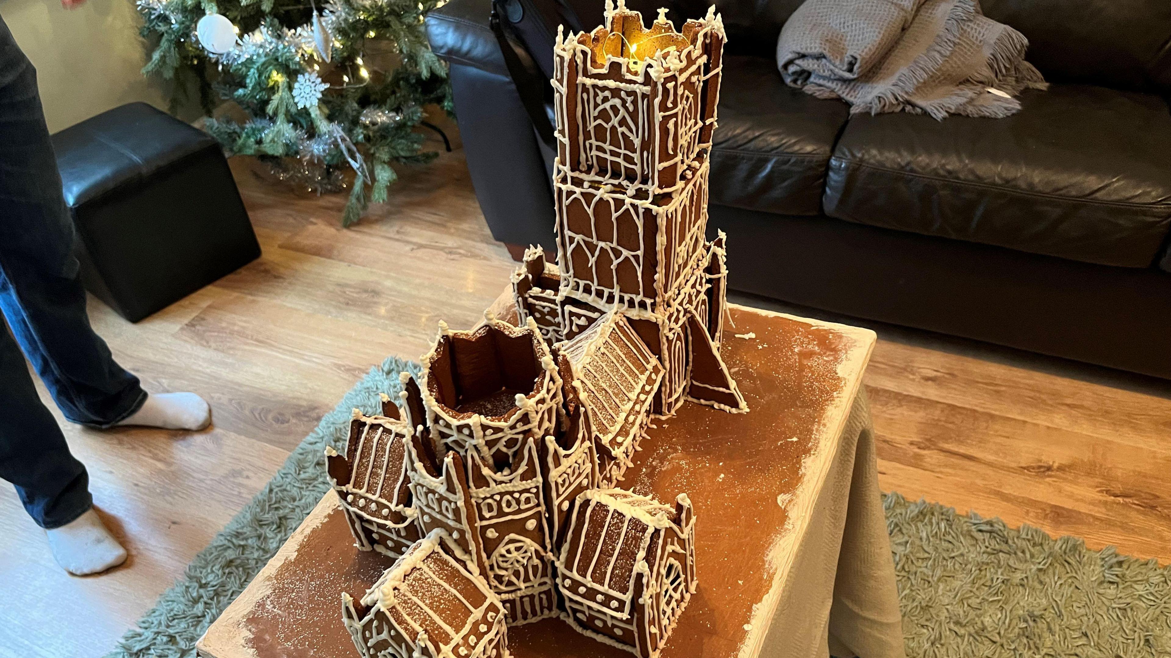 Gingerbread model of Ely Cathedral sitting in a living room, with a Christmas tree and decorations seen in the background as well as leather sofa and chair.