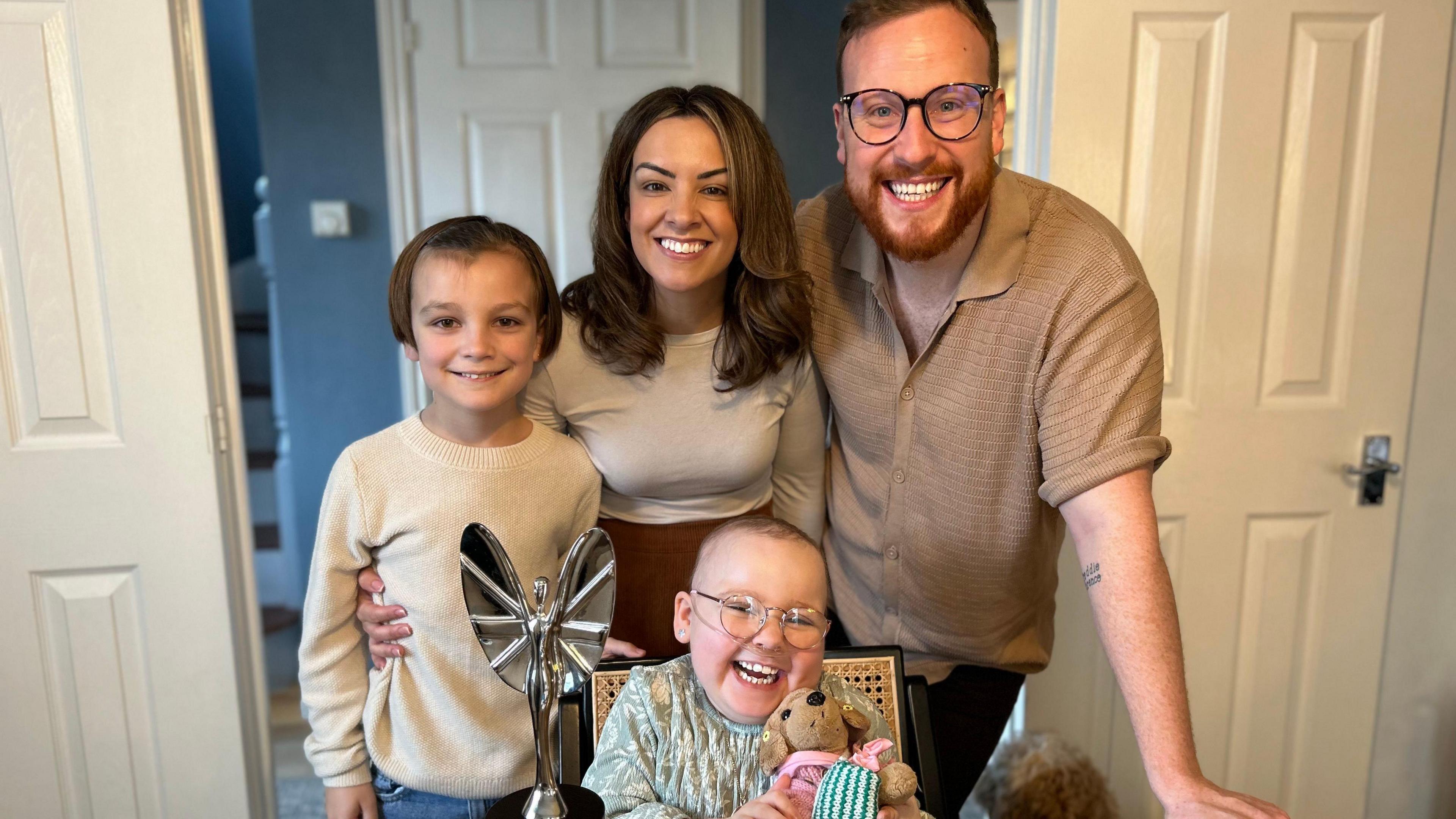 The bark family posing with Florrie's Pride of Britain award. Freddie, Florrie's brother who is 11 is on the left and wearing a cream coloured jumper. Mum Stacey Bark and dad Andy Bark are linking arms and grinning while Florrie sits at the table in front of them with a teddy. 