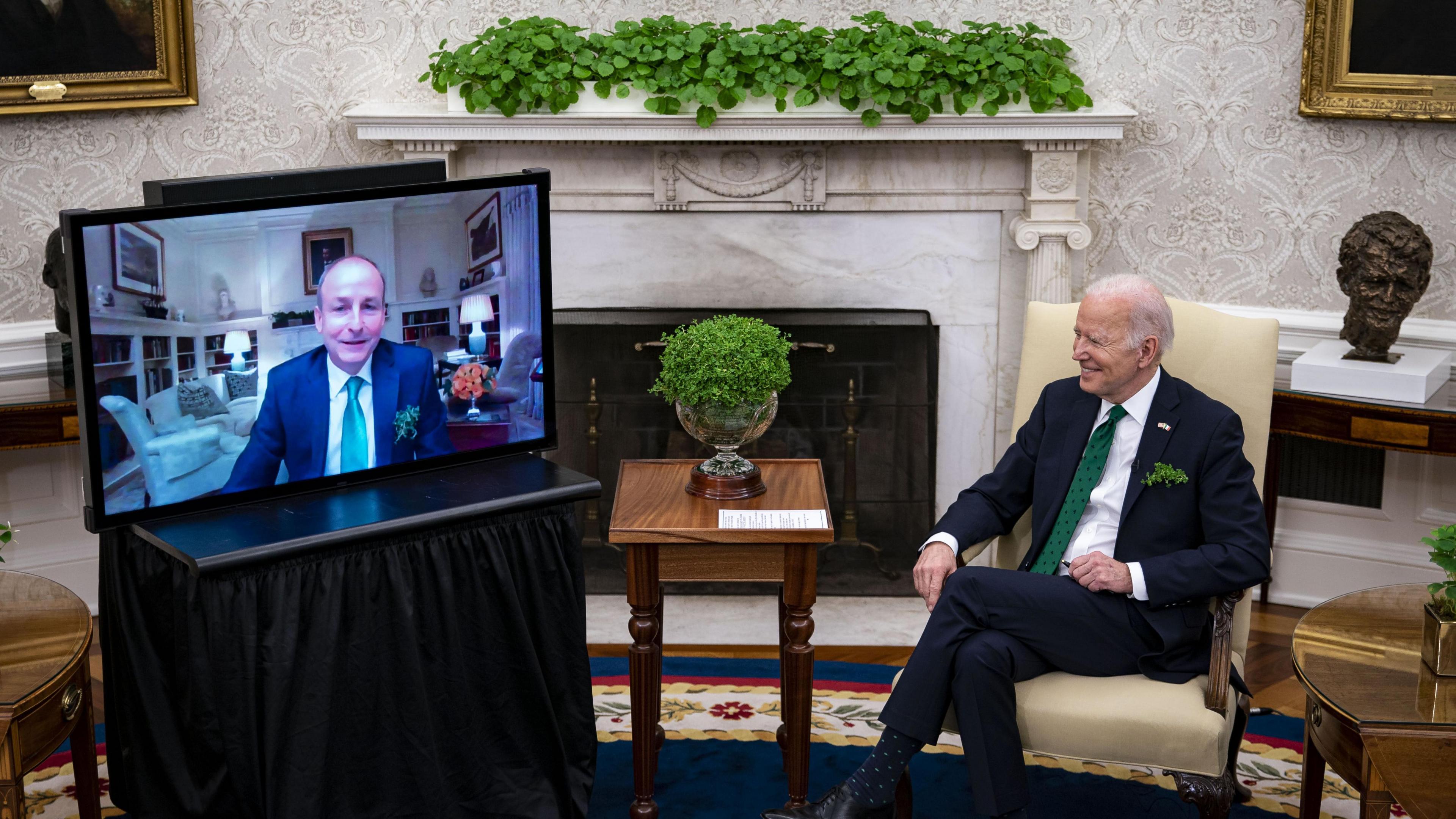 Micheál Martin pictured on a TV screen in the White House during a video call with former US President Joe Biden in March 2022.  Both men are wearing navy suits, white shirts and green ties.  A bowl of shamrock is on a table in front of Biden. 