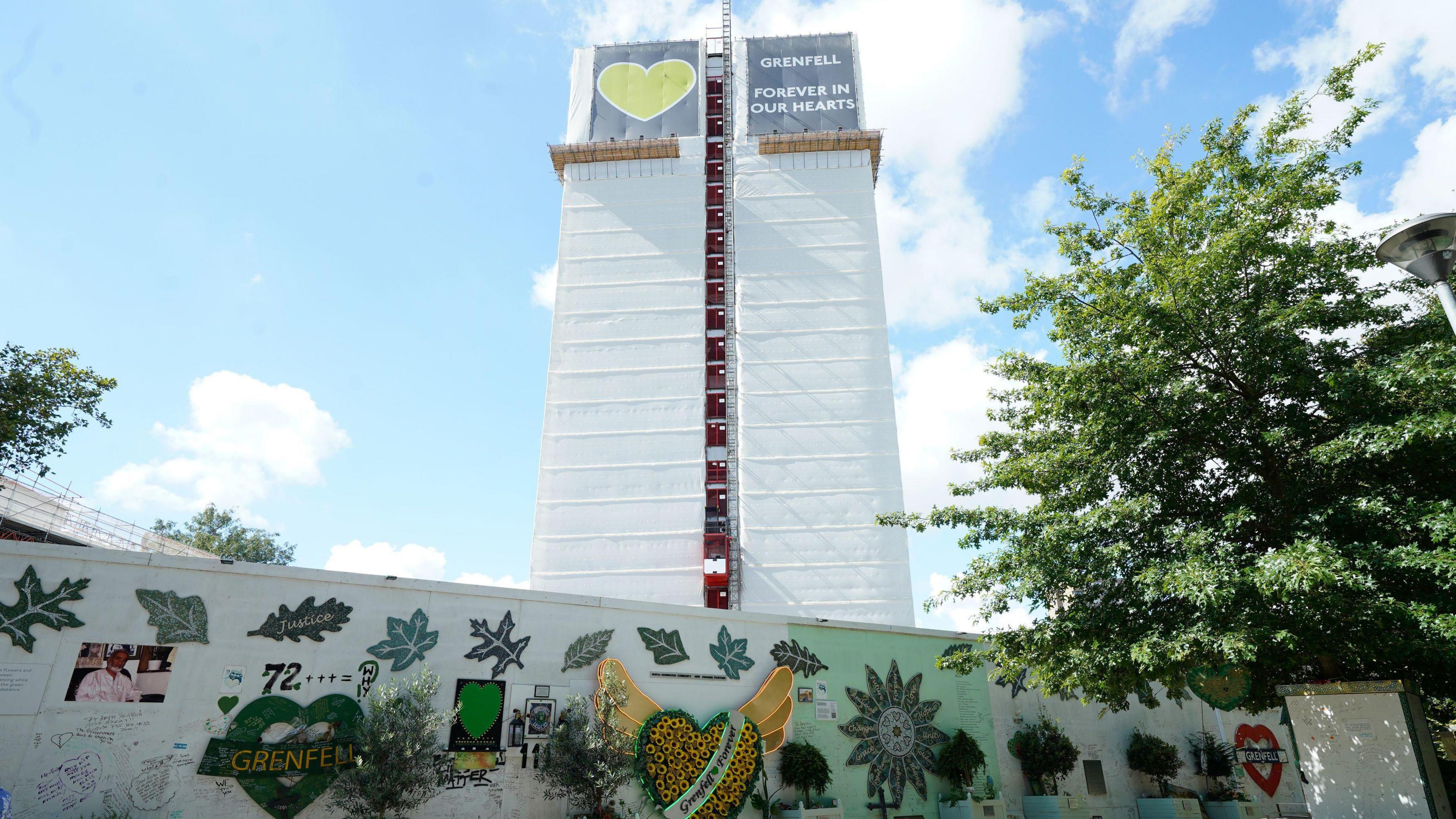 The Grenfell Tower block is wrapped in white plastic, with a large banner at the top reading Grenfell Forever in Our Hearts, with a large green heart. In the foreground is the memorial wall, covered in tributes to the people who died in the fire.