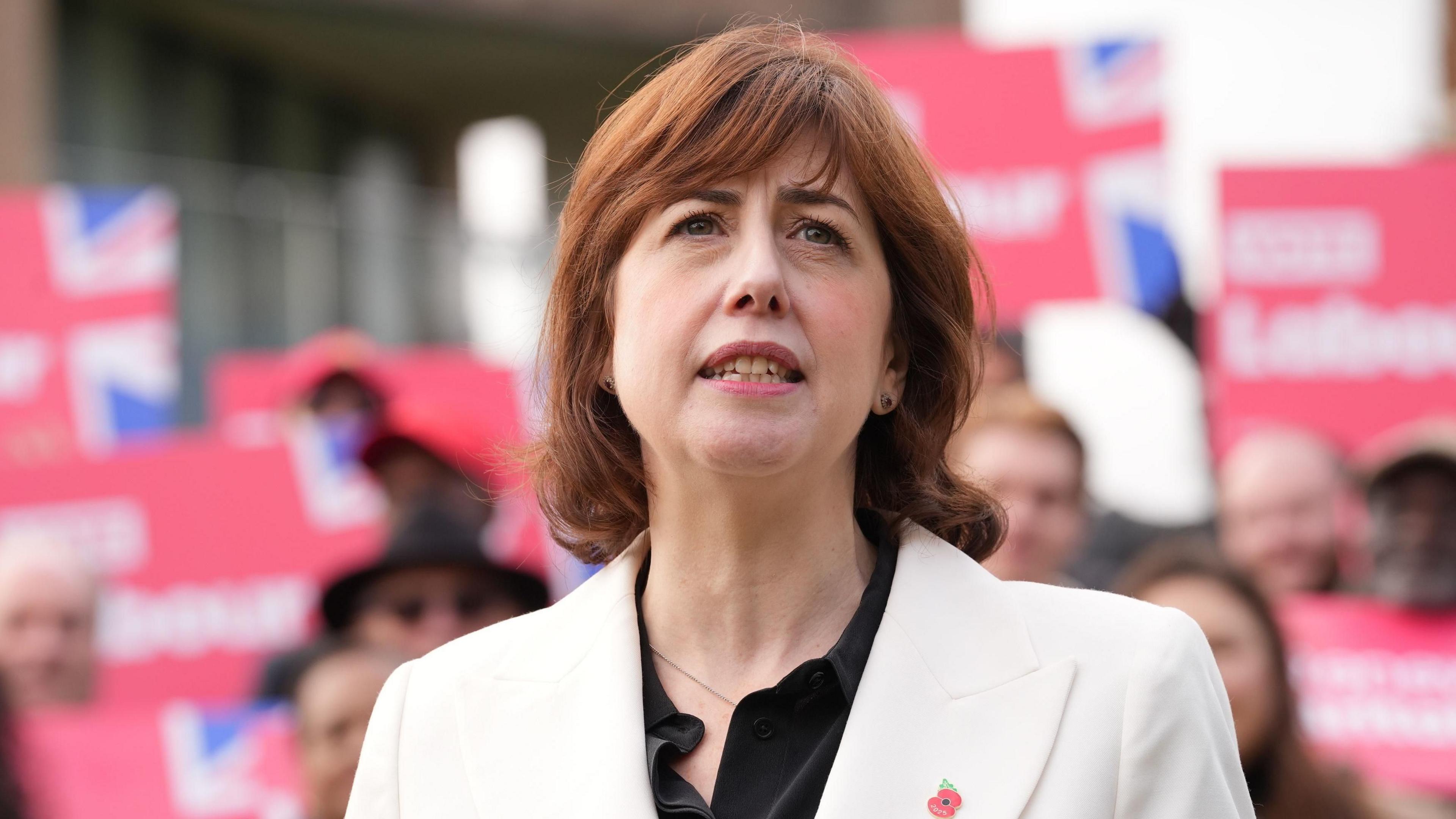 Lucy Powell wearing a white blazer and black top speaking in front of a band of supporters carrying red Labour slogans with union jack flags