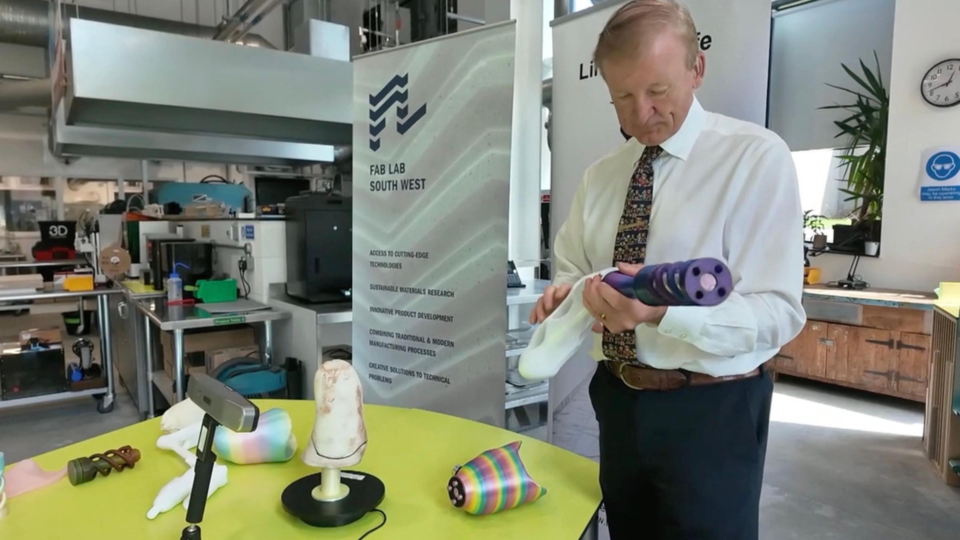 A picture of Mark wearing a white suit, with black trousers and a tie. He is holding a prosthetic limb and is stood in front of a green table. Behind him is an office with a number of items of machinery
