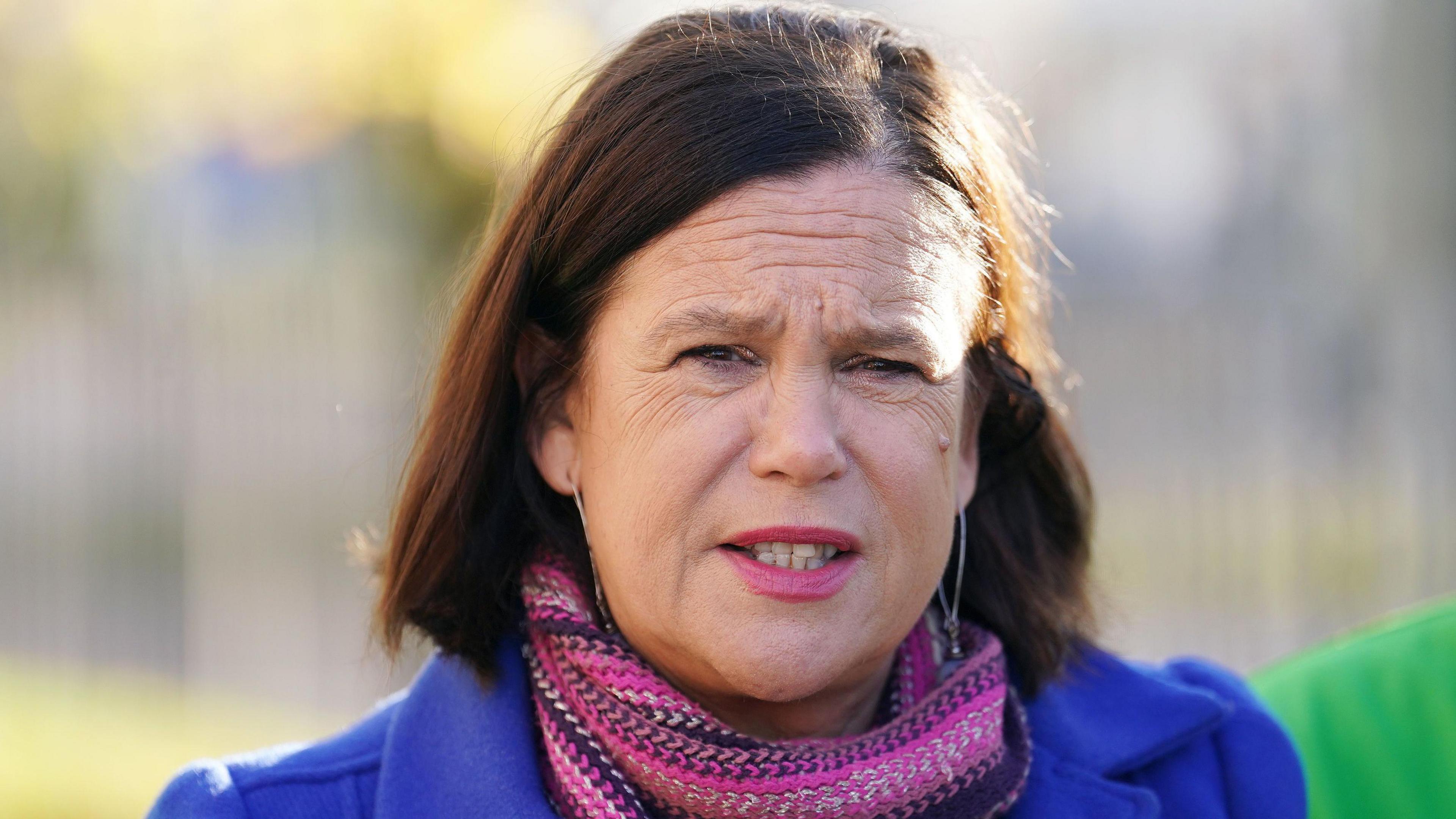 Mary Lou McDonald looking straight at the camera. She is wearing a blue coat and pink scarf. She had dark brown hair and pink lipstick.