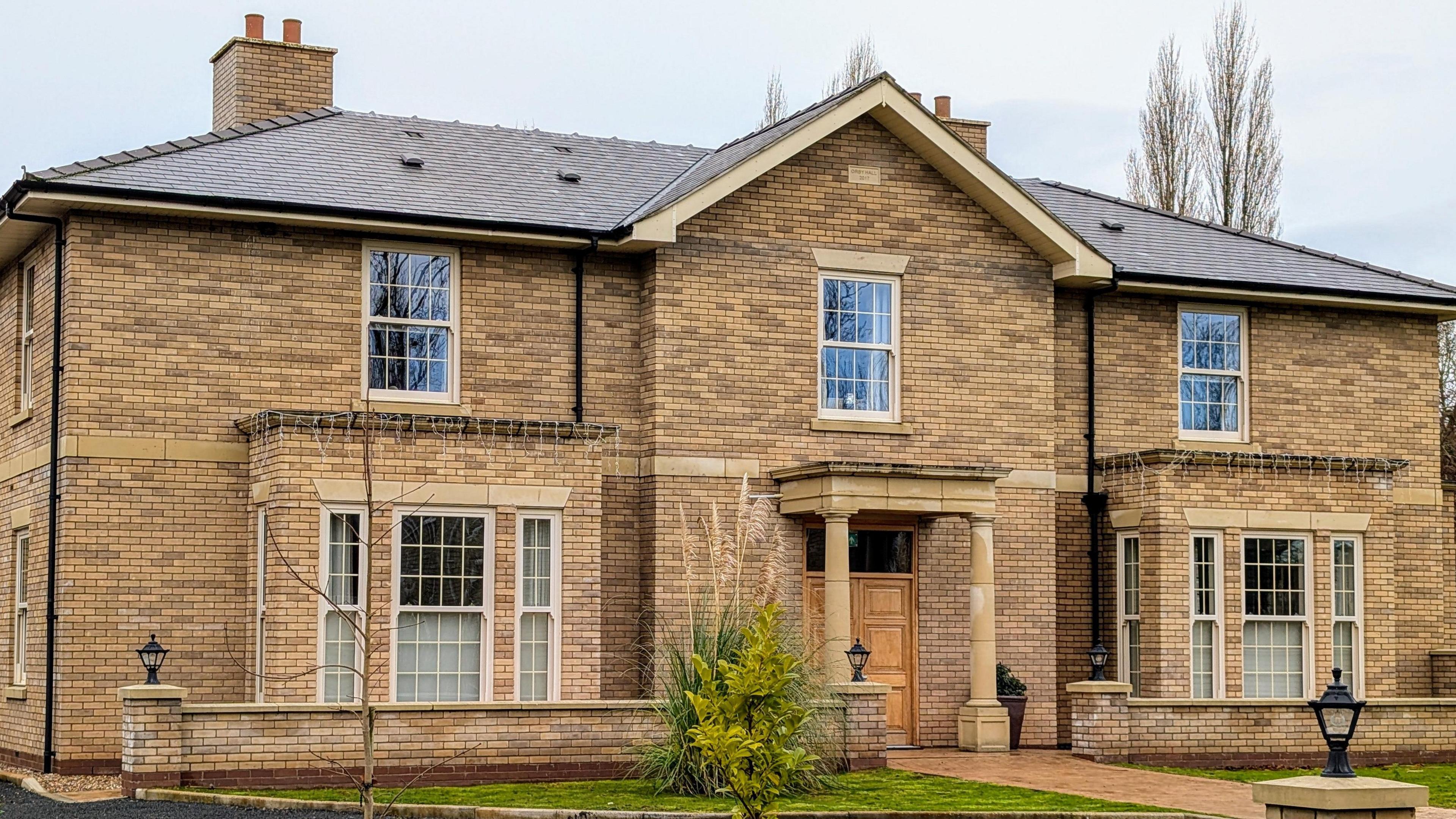 The Orby care facility, which is double fronted and built of light brick buildings. There are shrubs and grass verges in the photo.