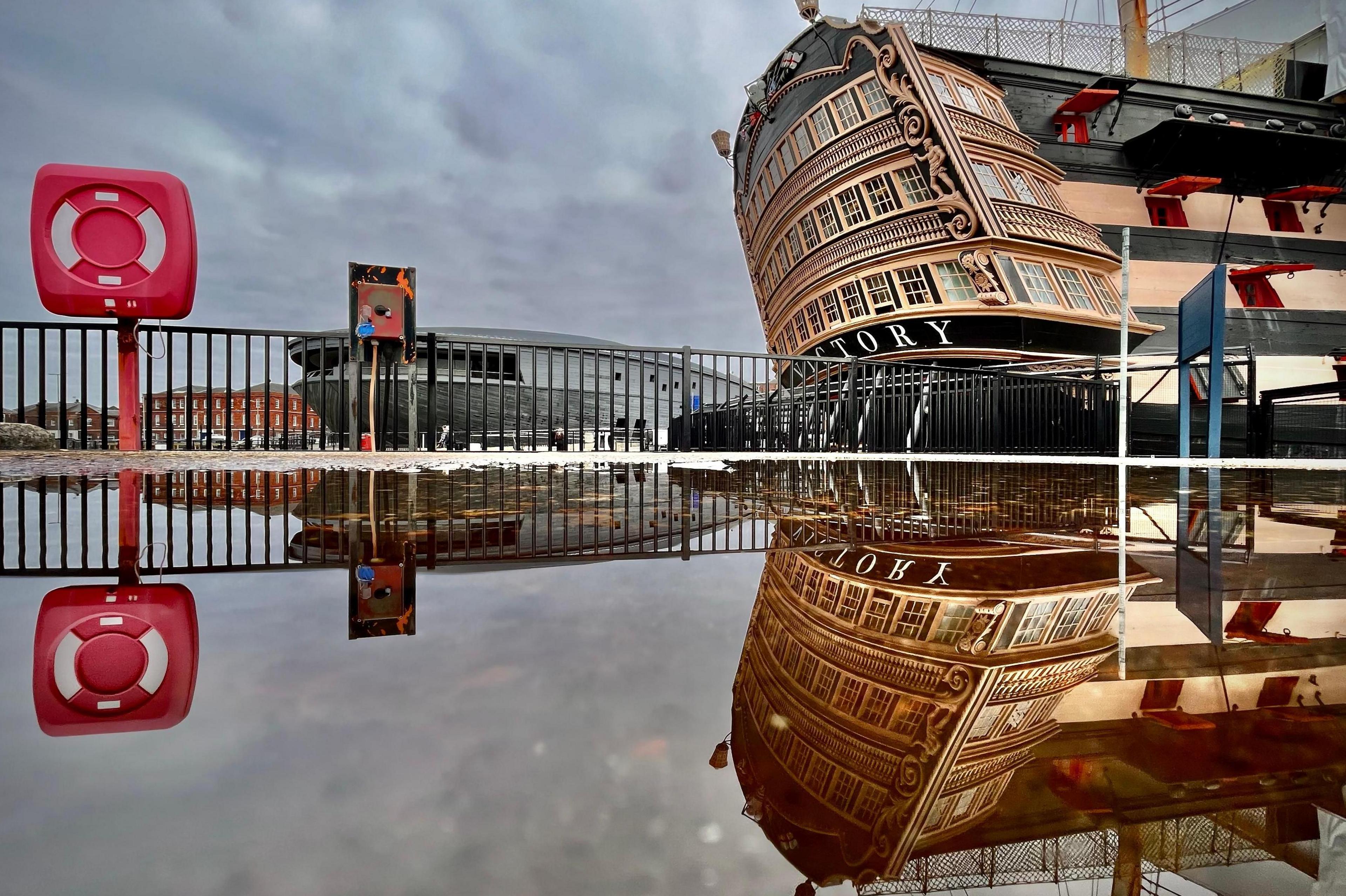 An image of the stern of the HMS Victory in its dock with a perfect reflection in water. A life ring in its housing is on the left of the shot, equally reflected. Railings and a building sit in the background.