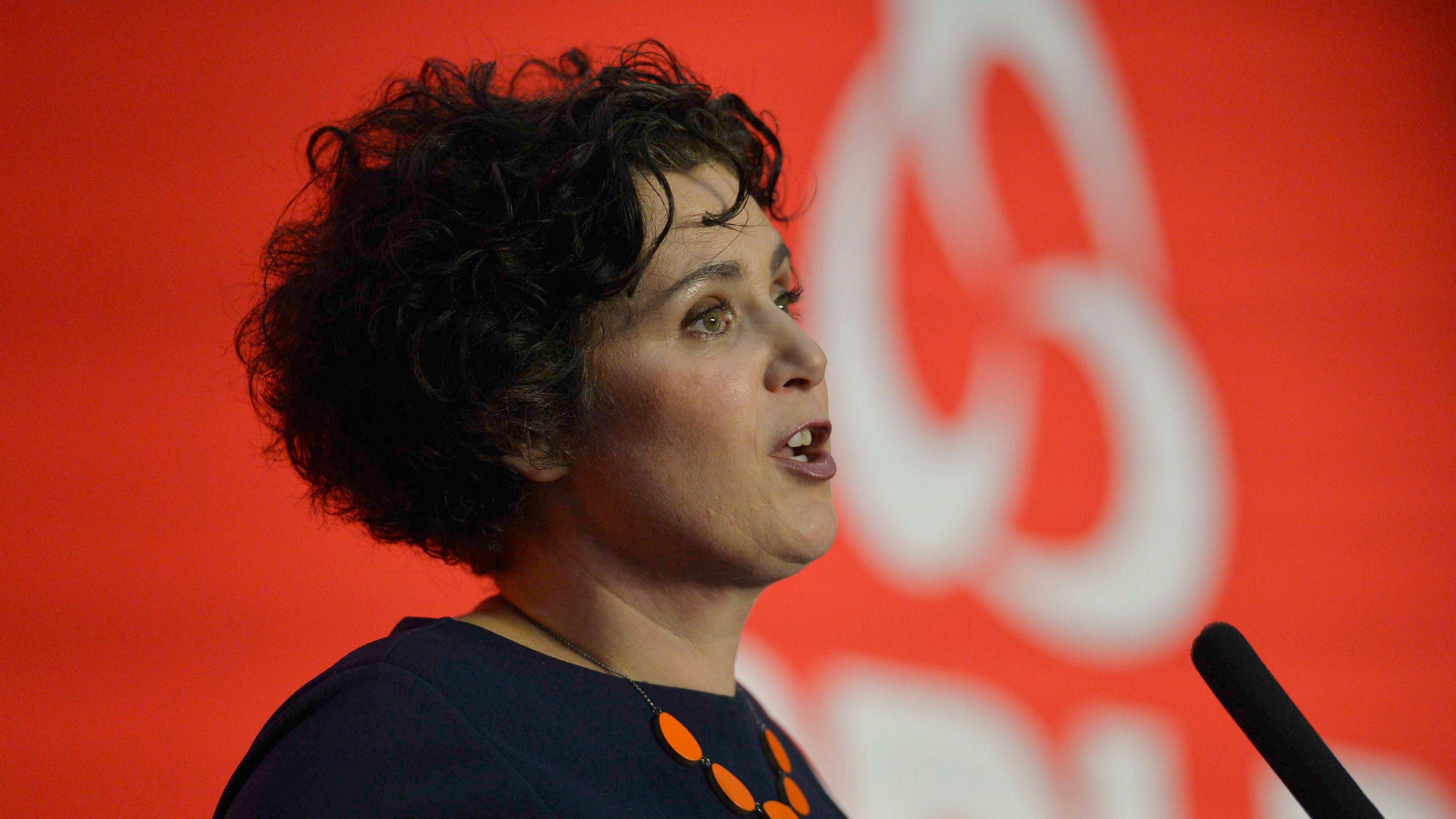 Claire Hanna. She has black curly hair, wearing a navy top and orange necklace. She is standing in front of a microphone.