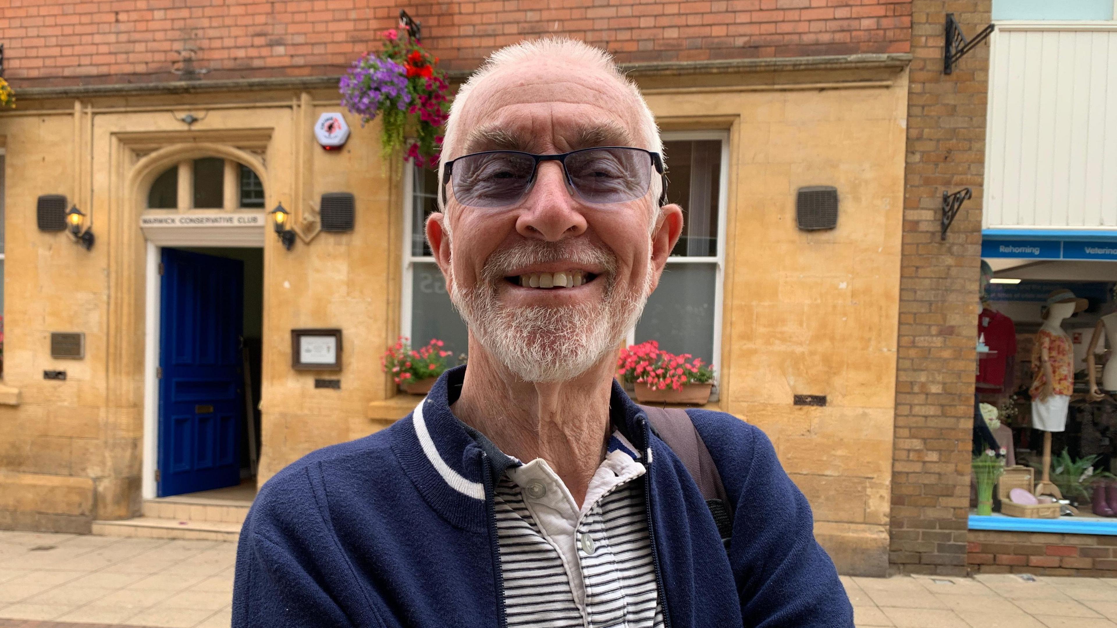 Vic Mann smiles at the camera. He is wearing a blue cardigan over a white and blue stripped polo shirt. He has glasses, white hair and a white beard.