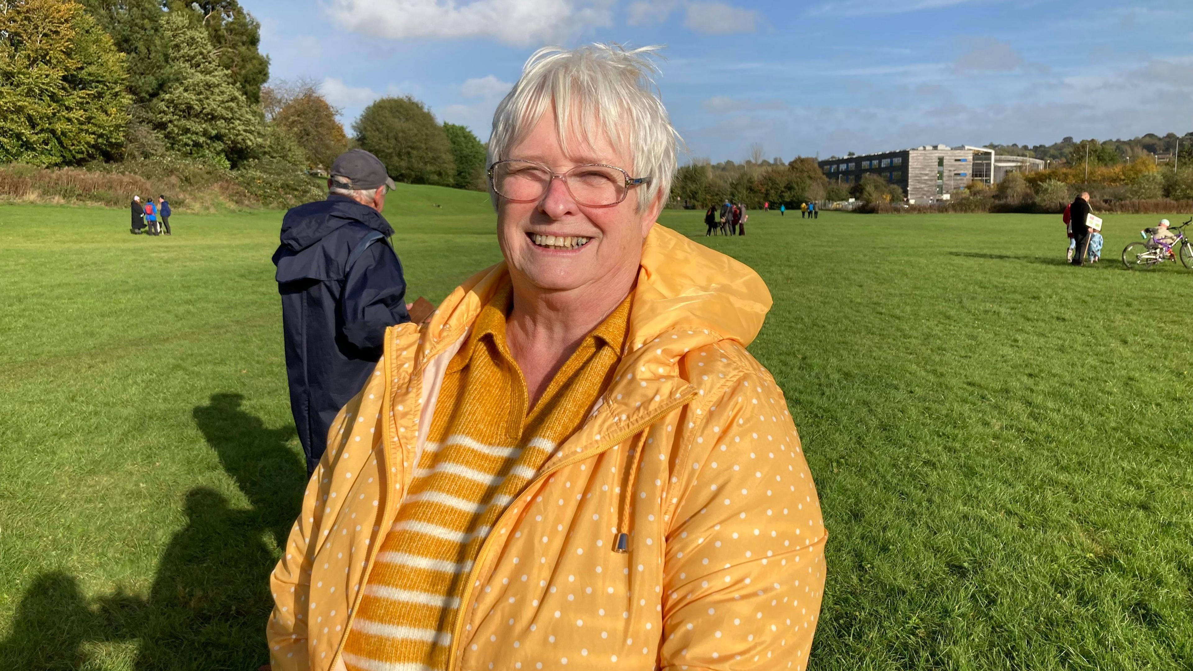 Liz Scott with short white hair and glasses wearing a yellow raincoat and standing on grass