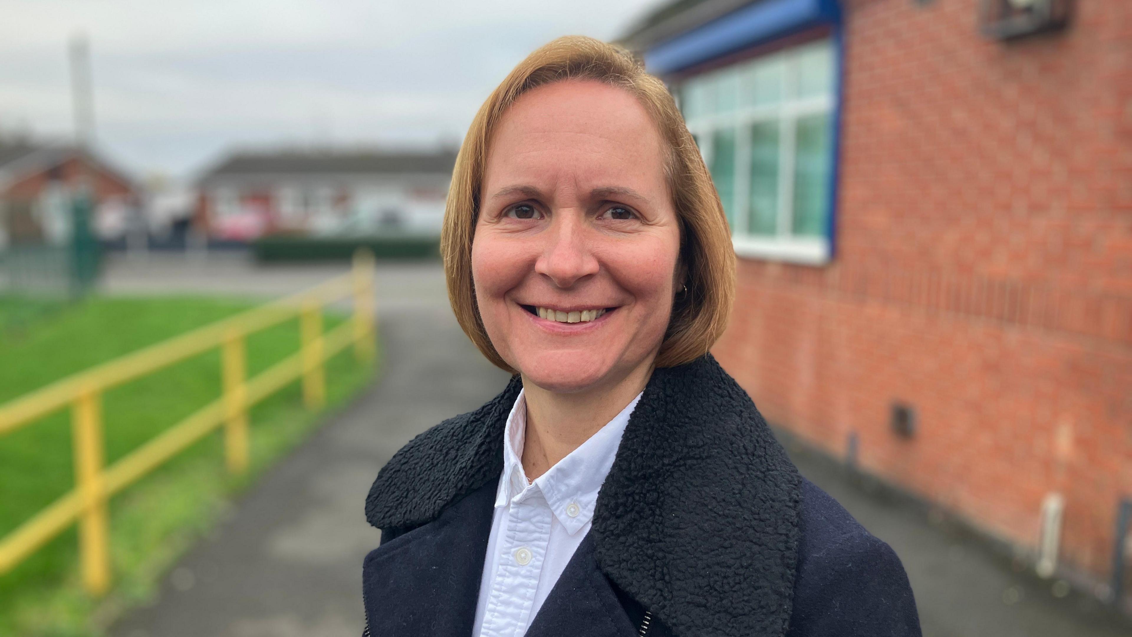 Woman with short blonde hair, wearing a blue shirt and navy coat with a thick black collar, smiling with a yellow fence and brick wall behind her