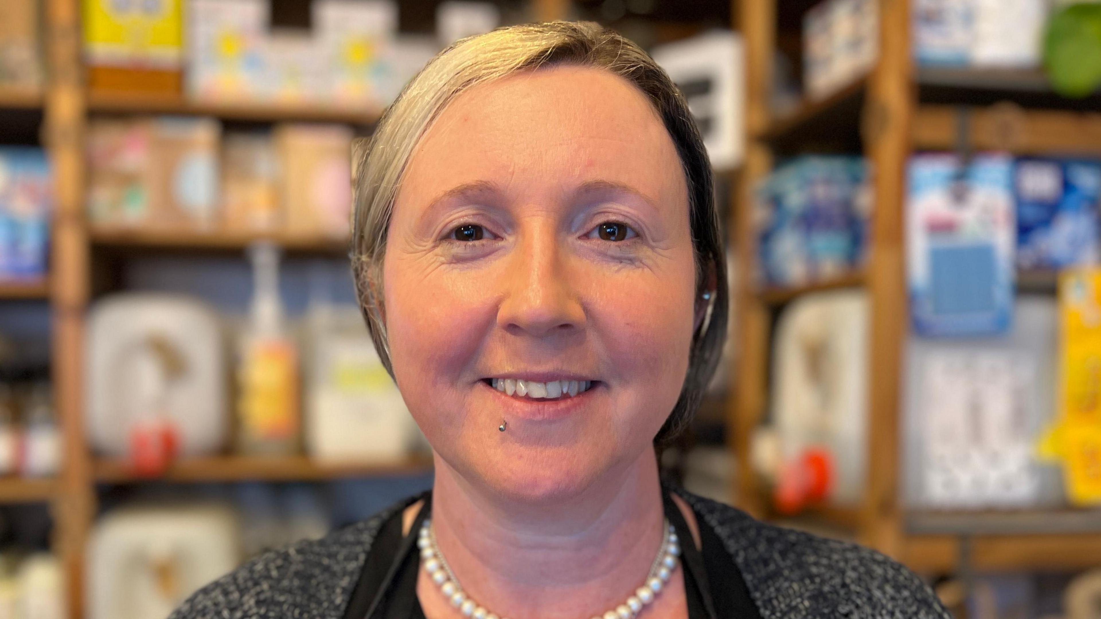 Johanne Bridges is smiling, she has a piercing below her lips and brown eyes. Her hair is blonde on one side and browner on the other. She is wearing a pearl necklace and a grey cardigan with a black apron over the top.