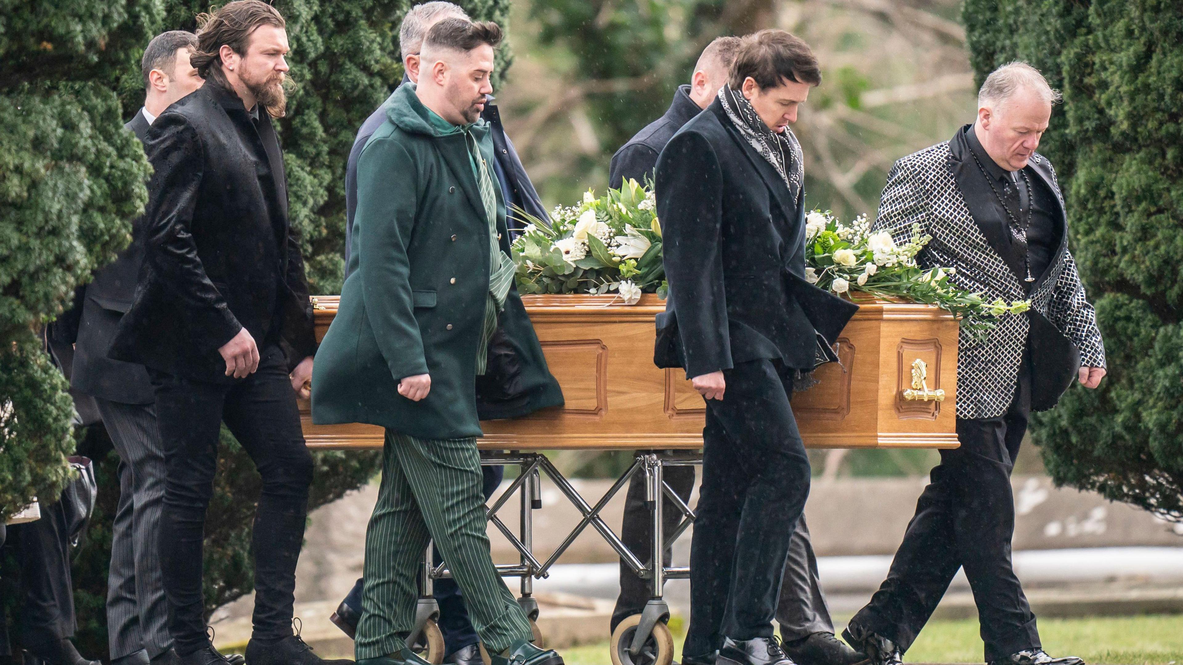 A light brown wooden coffin adorned with white flowers is accompanied by mourners.