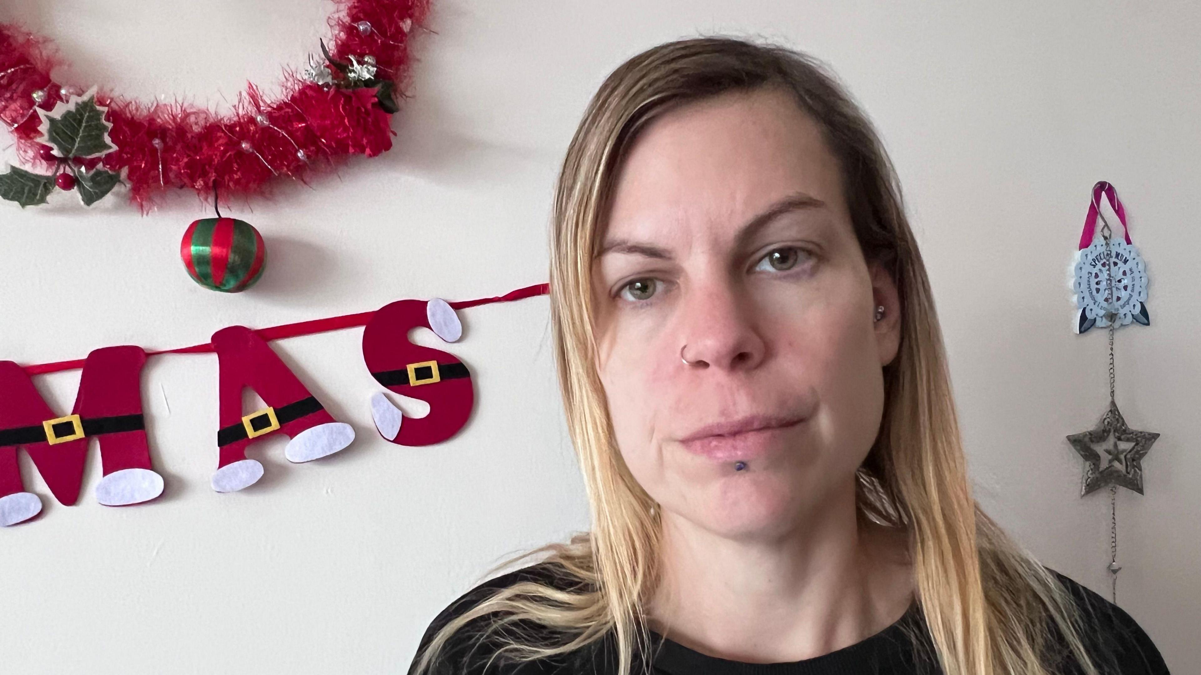 A woman with long straight blond hair wearing a black jumper, behind her is some Christmas decorations. She is looking straight faced.