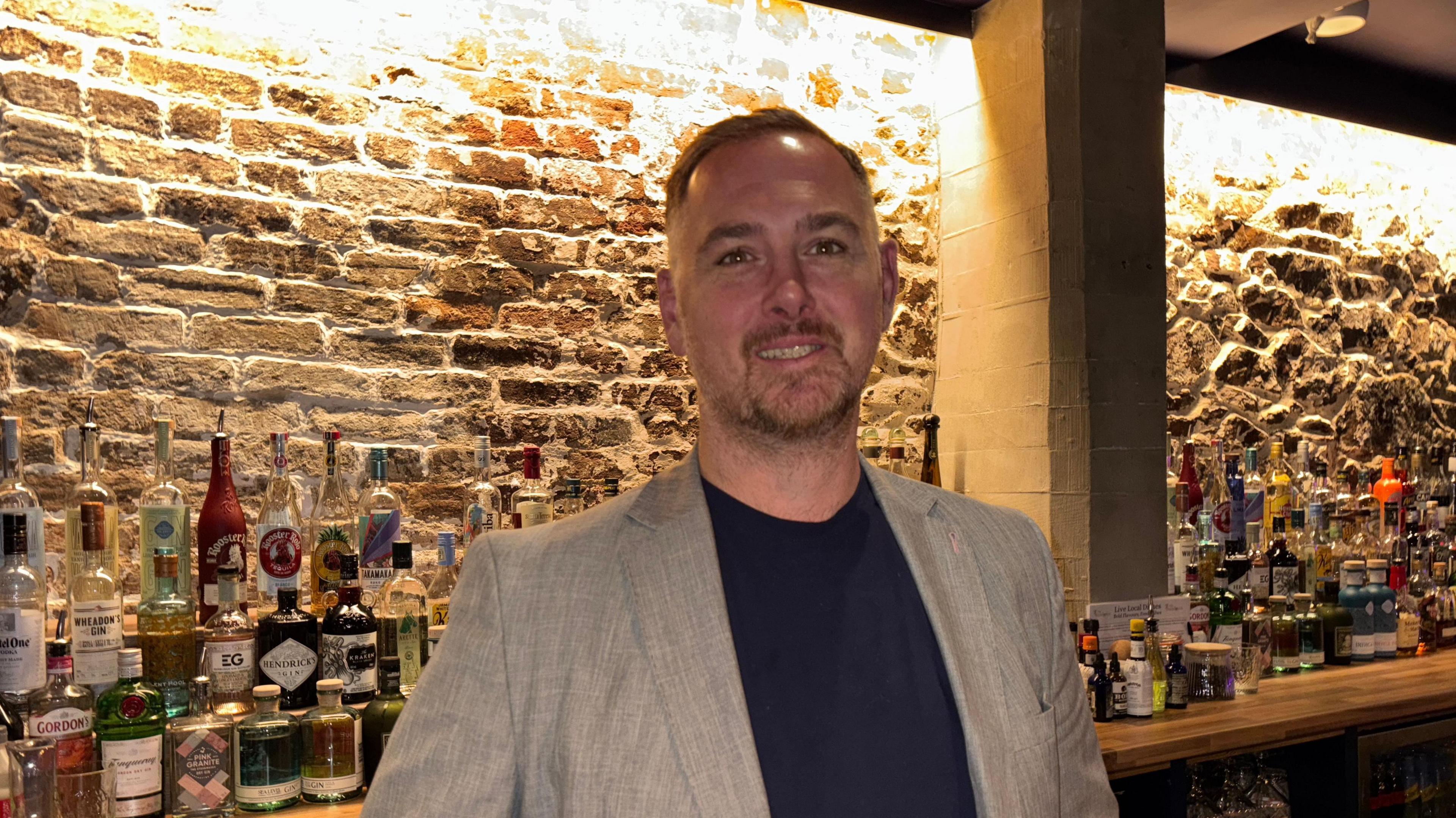 Simon Soar, a man with short dark hair and a goatee-style beard. He is wearing a beige suit over a dark blue, round-necked shirt. Behind him is a number of bottles of spirits, arranged along a bar. Behind the bar is an exposed brick wall.