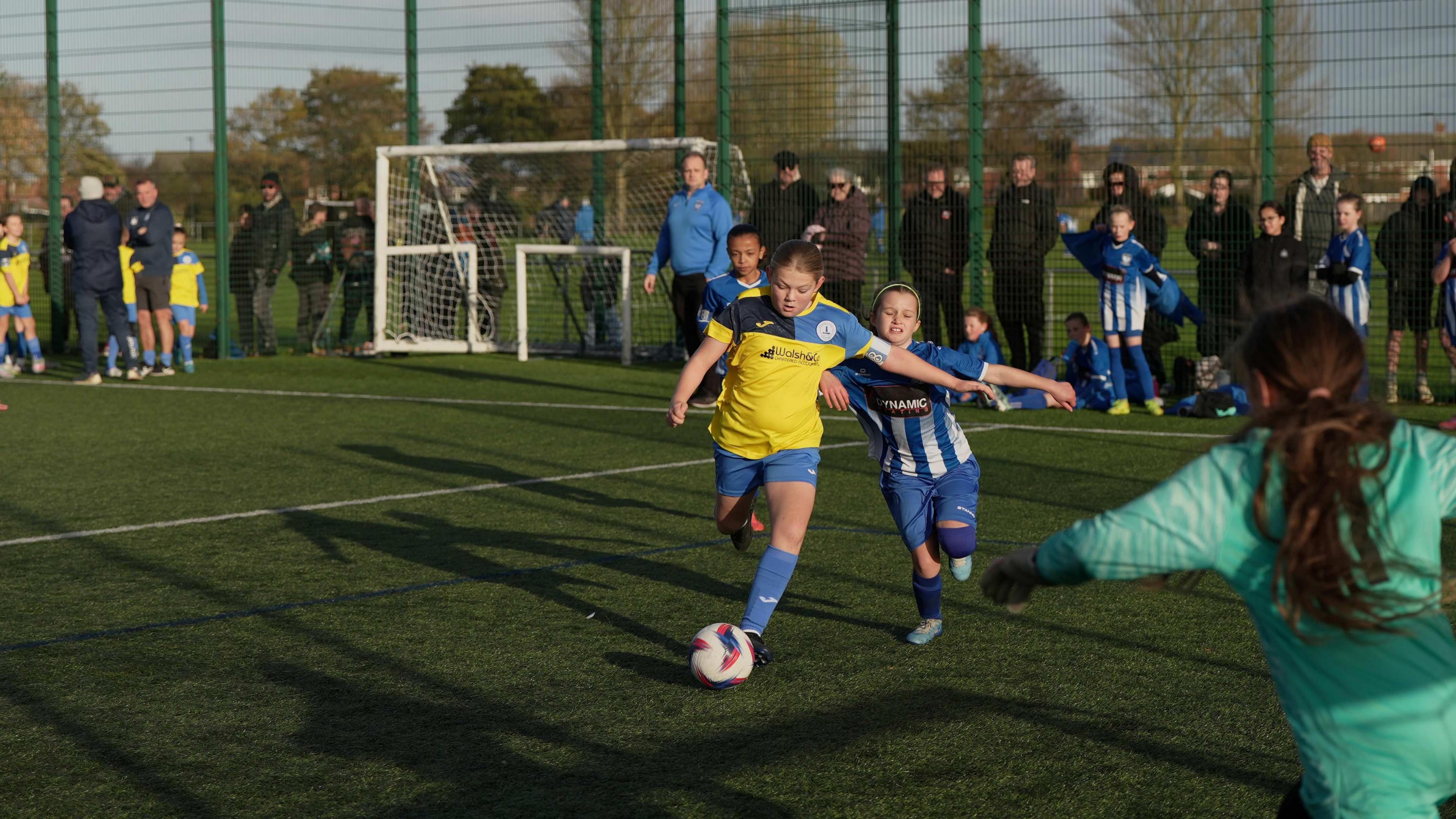 Two girls in Whitley Bay Football Club and Whitley Bay Sporting Club kit play football with each other on an artificial turf pitch.