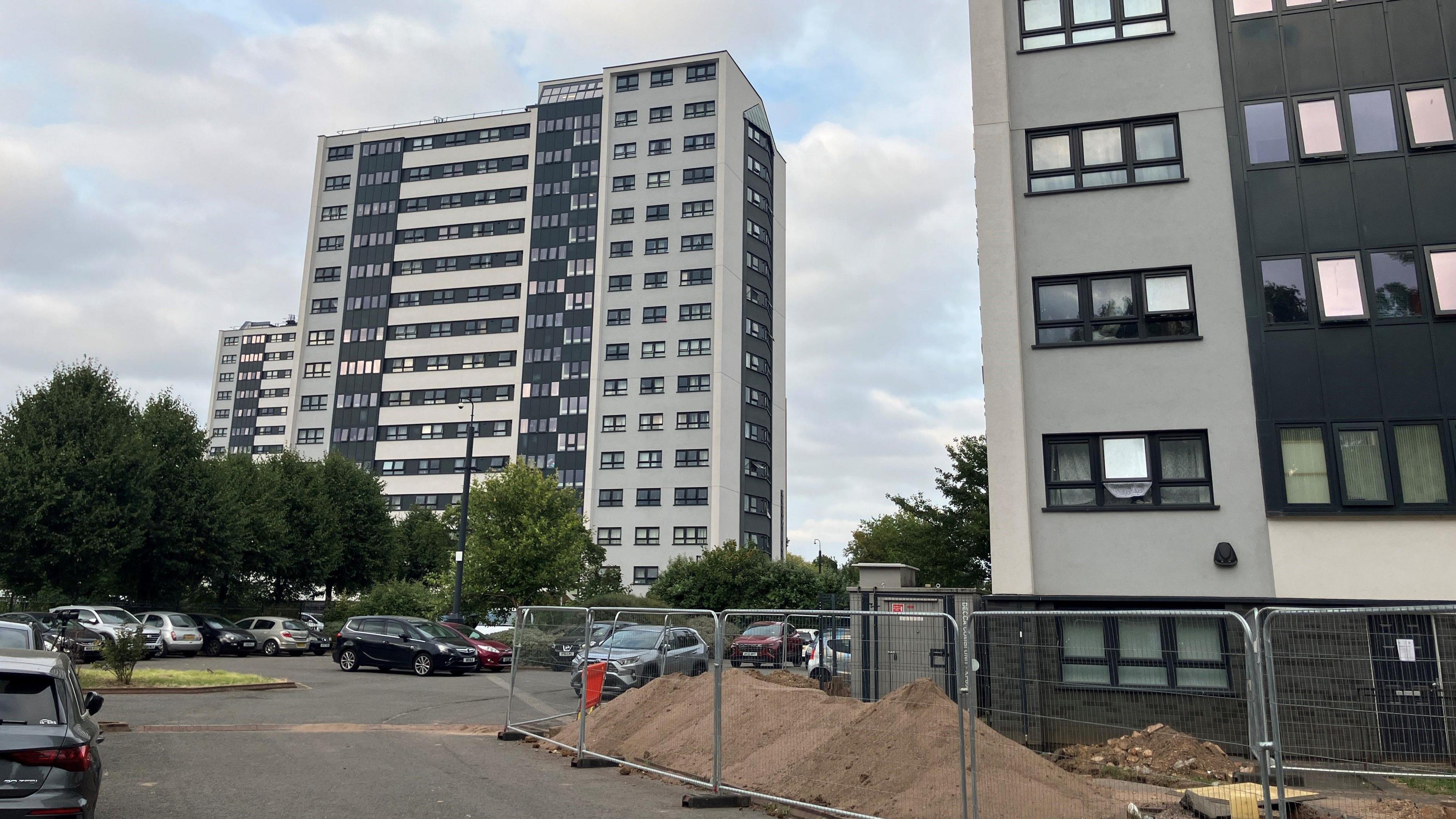 A row of three 15-storey tower blocks, with one fully visible in the middle of the shot, the side of one block in the foreground, and the third visible in the distance behind the middle one. All three are rendered in light grey with black panels and windows.   