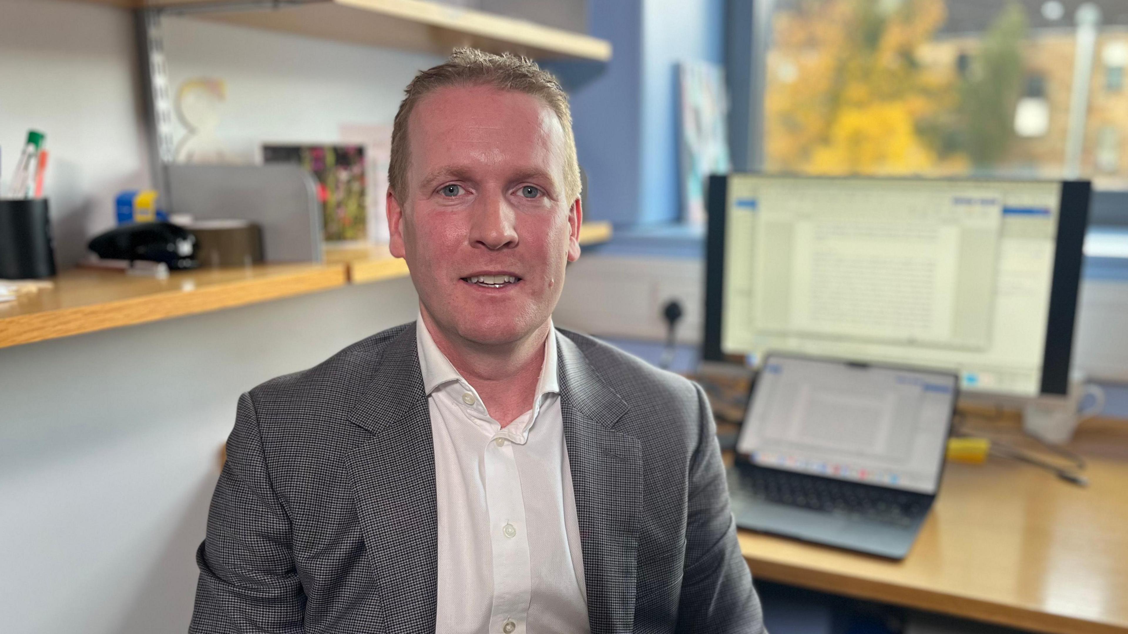 Prof Millar is sat in his office with a laptop and a computer monitor on a desk in the background. There is a bookshelf behind his head with various office accessories such as a stapler and pen holder. Prof Millar is sat looking at the camera. He has short sandy hair and is wearing a white shirt and a blazer jacket.