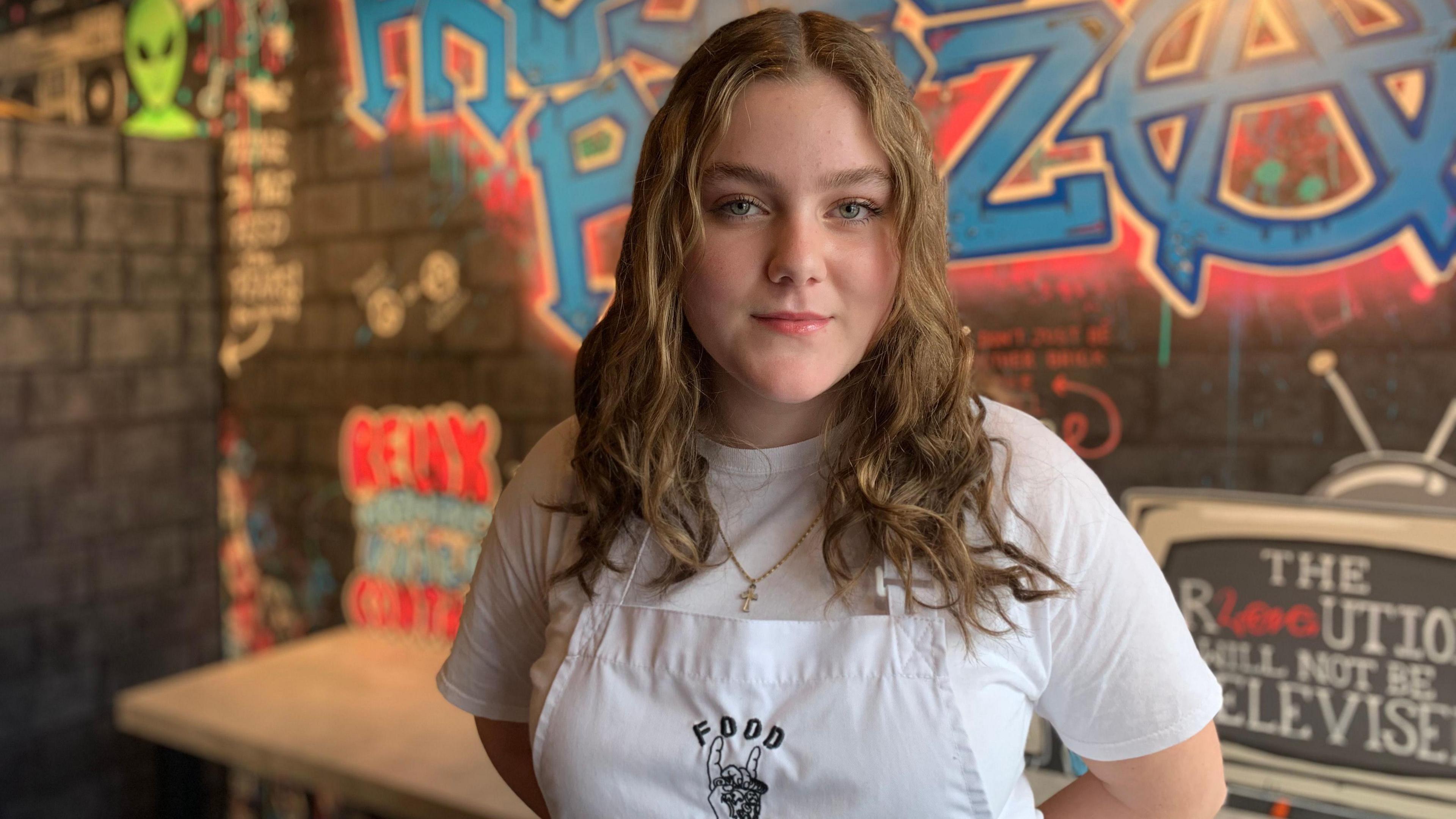 A long-haired young woman in an apron and a white t-shirt stands in front of a counter in a takeaway pizza restaurant