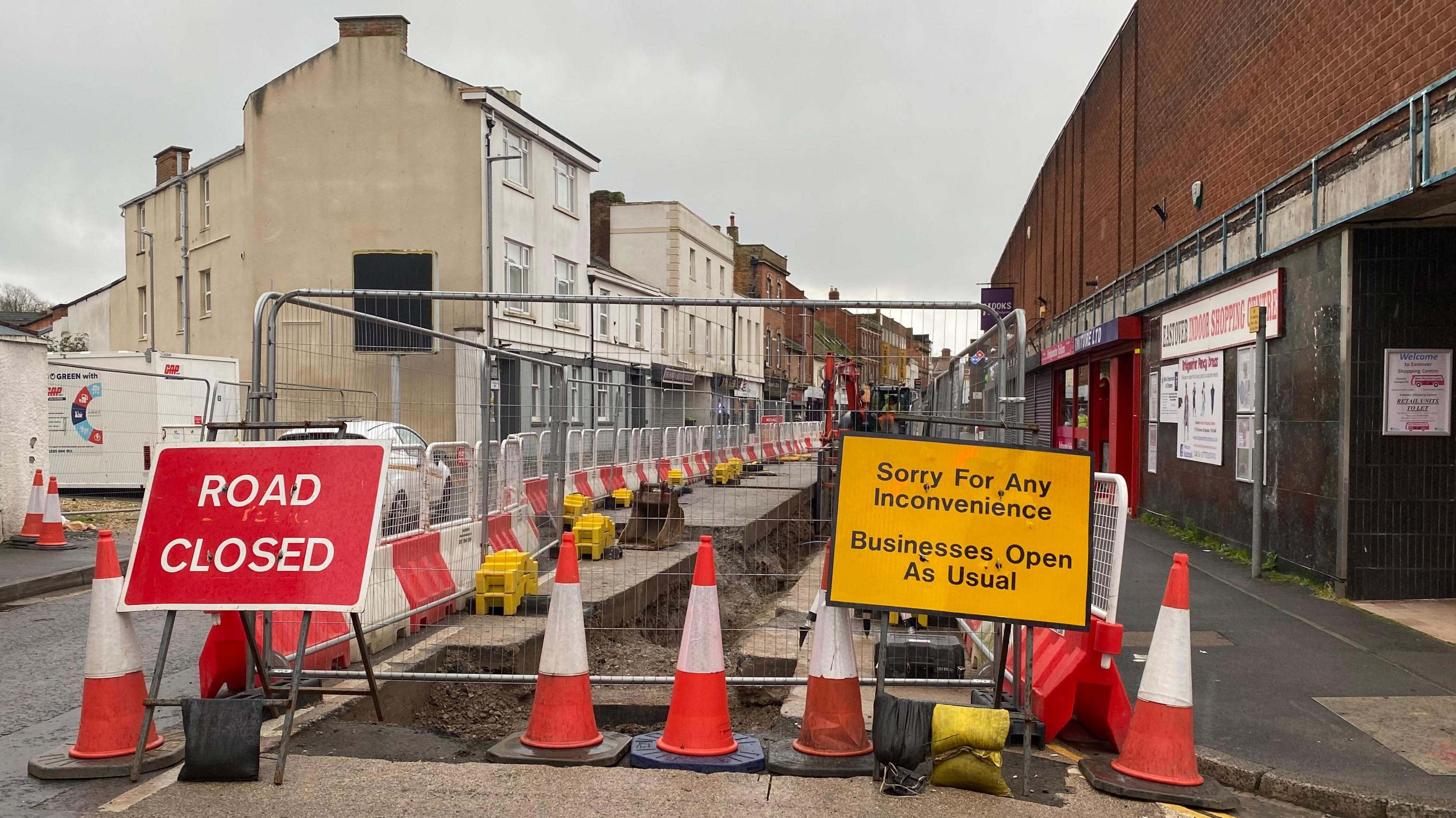 Fences block the road, with signs saying 'road closed' and 'sorry for any inconvenience - businesses open as usual' either side. There are traffic cones at the front of the fence, and the road is flanked by properties.