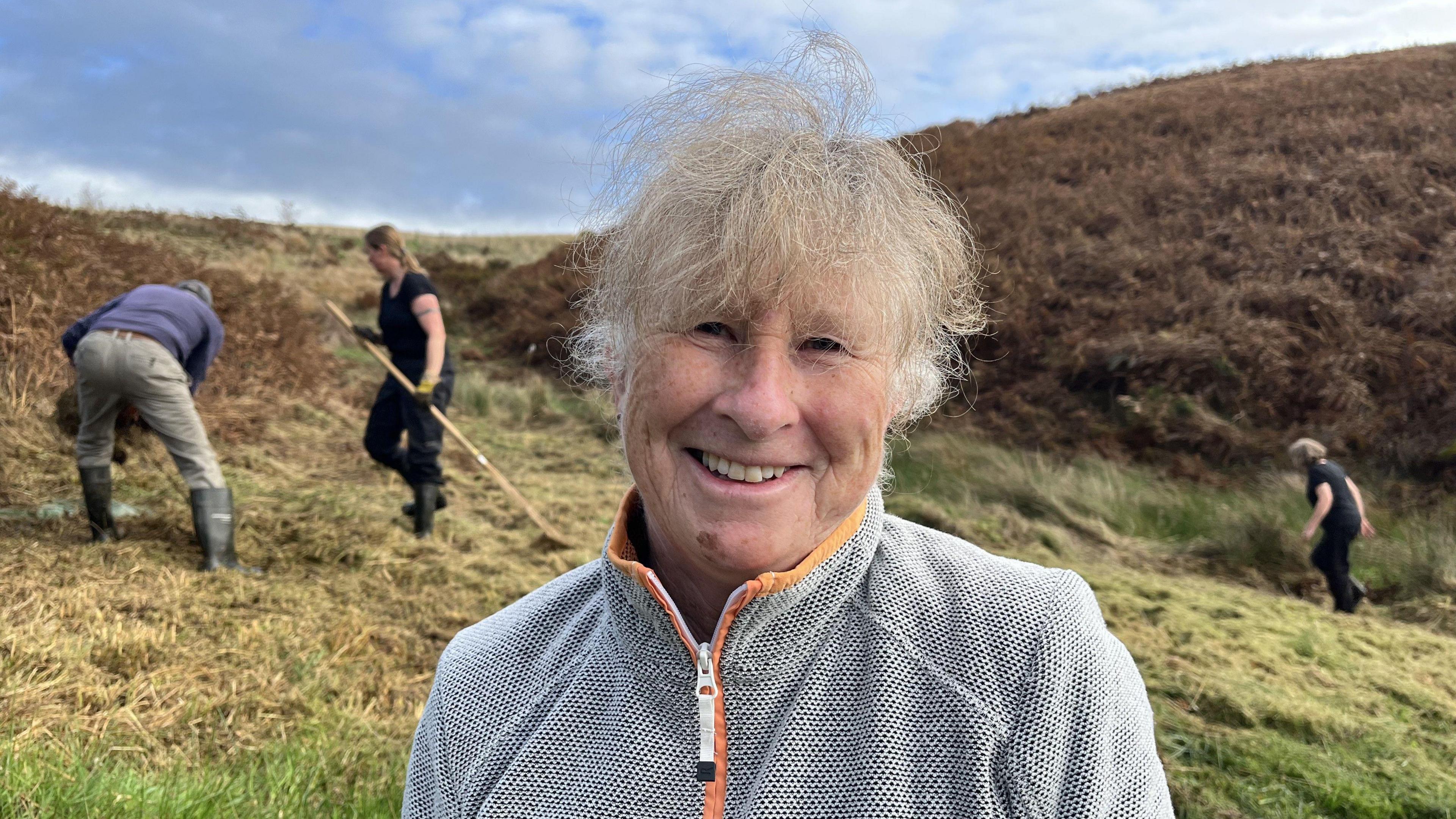 Jan is standing on a hill, she has a grey zip-up sweater on, and has short, blonde and grey hair. She is smiling at the camera. In the background are two volunteers who are raking grass