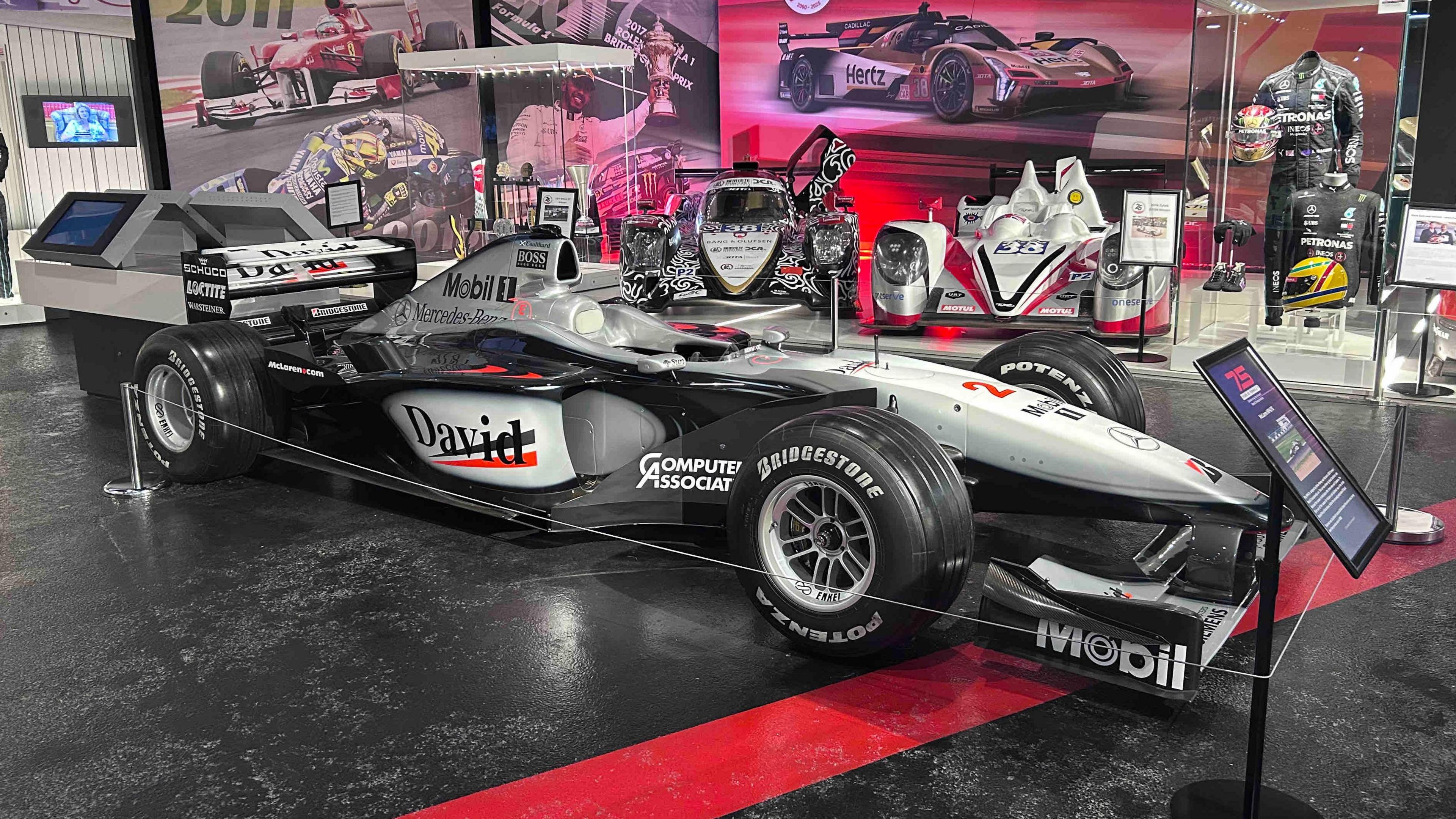 A black racing car with silver trim. The name David is seen in black writing on a white background on the side of the body, alongside "Computer Associates" and Mobil. There is an information stand in front of the car, which is parked on a black and red surface. There are display boards with pictures of racing cars behind it.