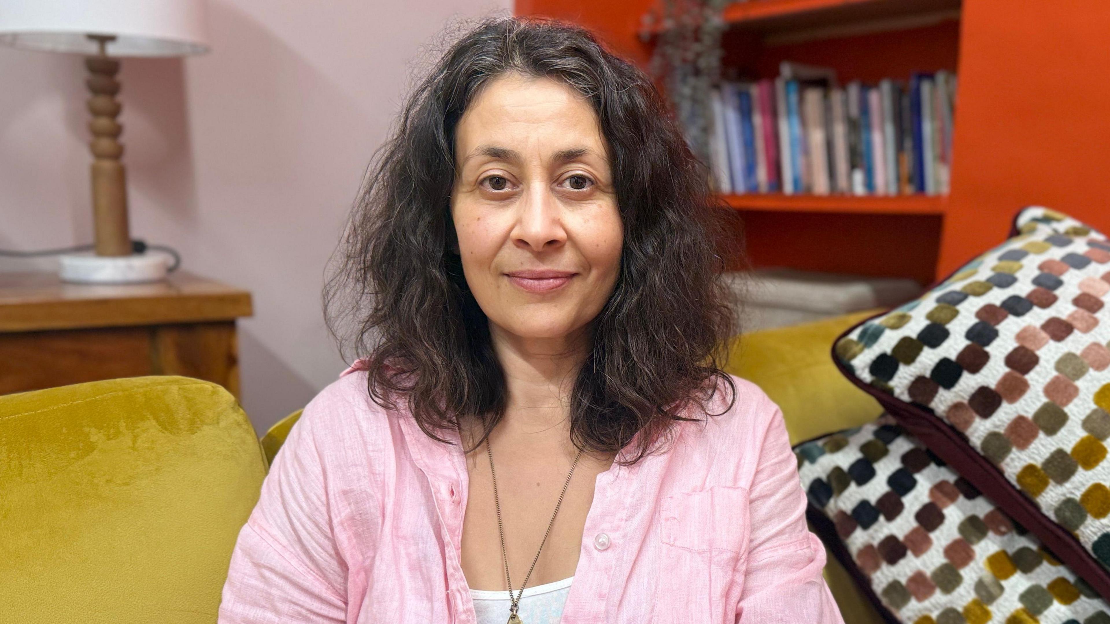 Rosie Lewis looks at the camera. She has shoulder length thick wavy hair and is wearing a bright pink shirt over a white t-shirt. She is sitting on a yellow sofa with a book case behind her.