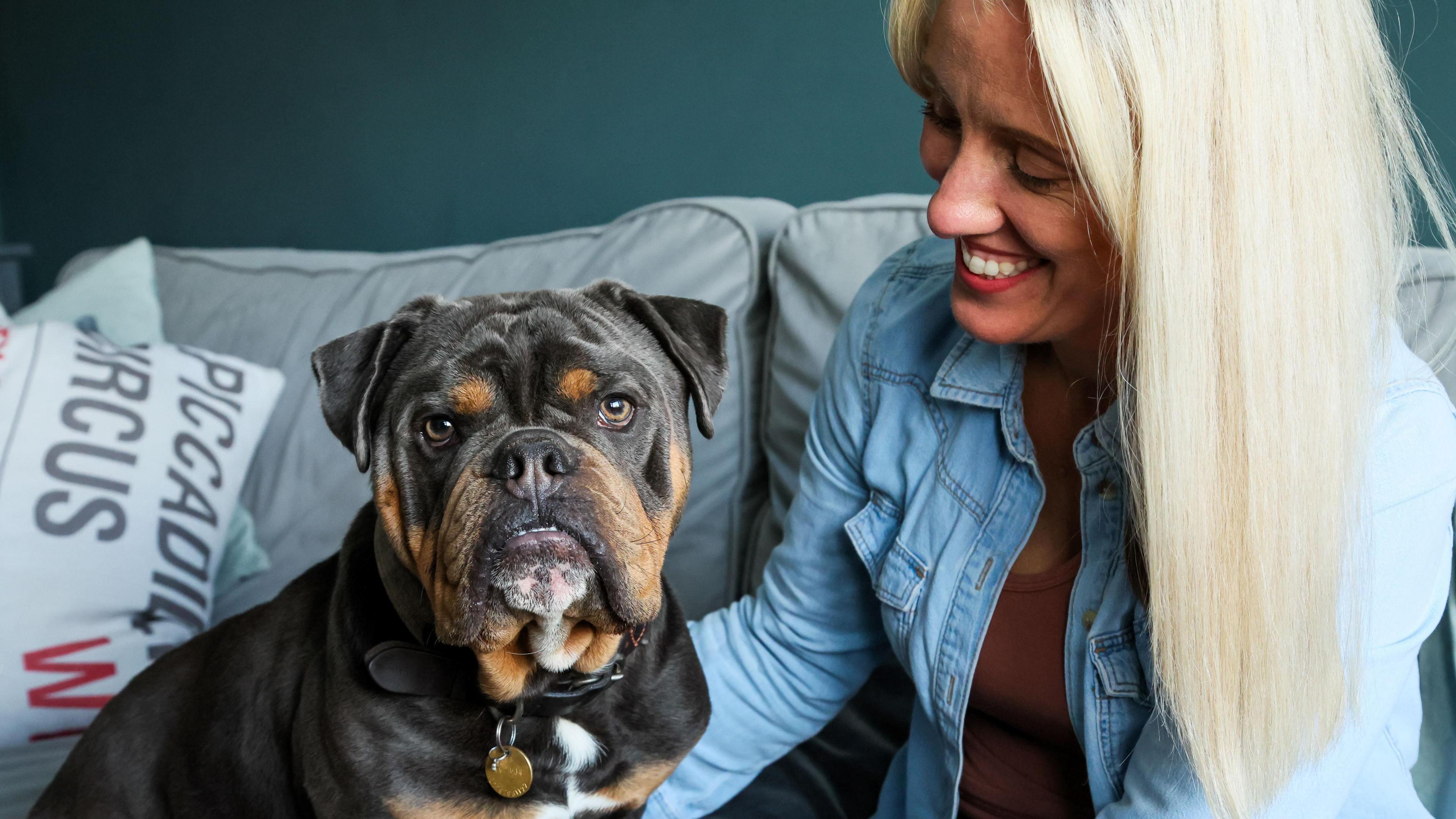 A woman with straight blonde hair and a denim shirt sits on a sofa, smiling down at a bulldog perched to the left who looks at the camera. A cushion saying 'Piccadilly Circus' is behind him.