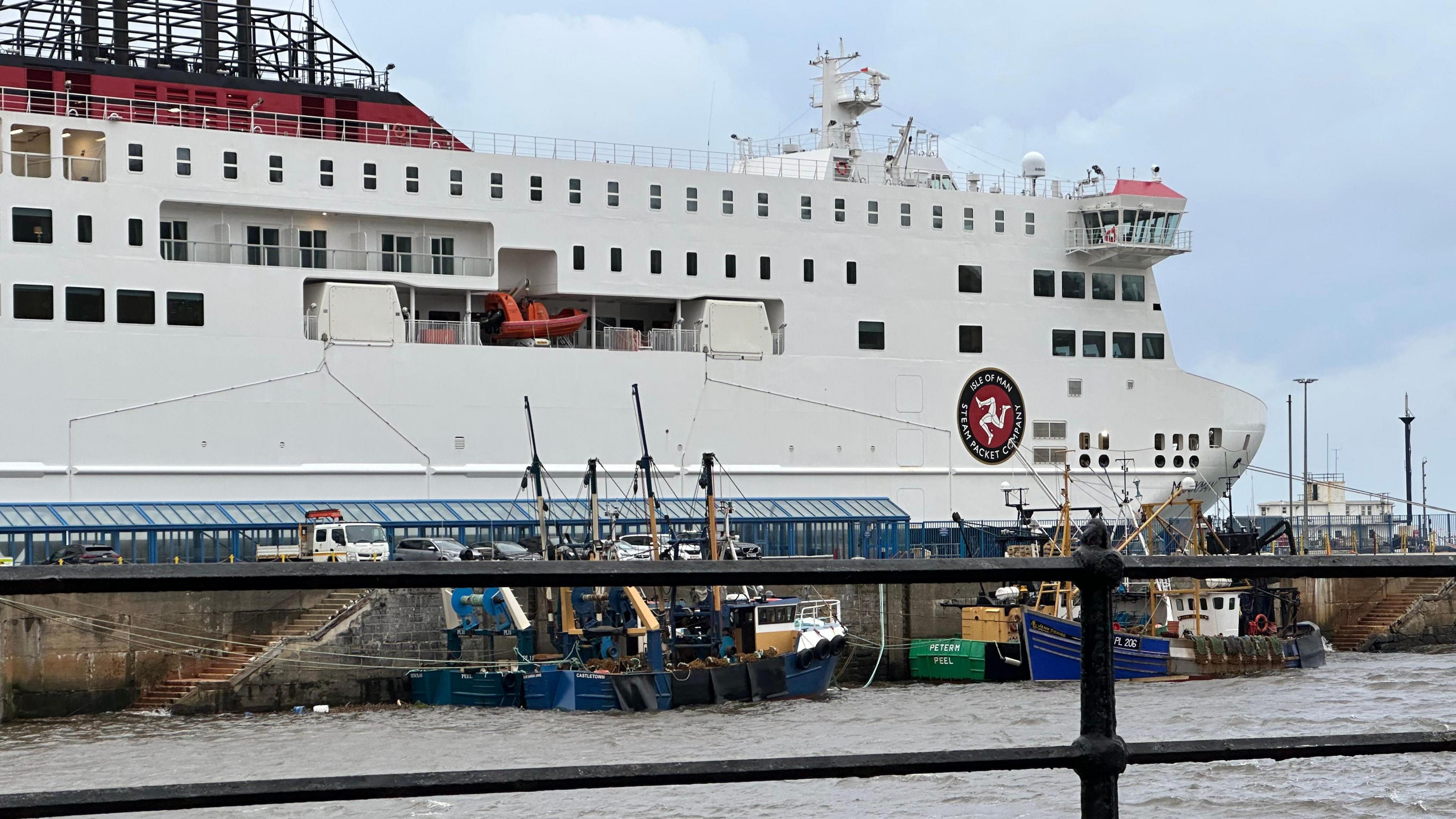 The bow of the Manxman moored in Douglas Harbour. The vessel is a large white ferry with the Isle of Man Steam Packet's logo featuring a three legs of Mann symbol in a red circle with a black band around it bearing the firm's name in it. Fishing boats can be seen tied up on the opposite side of the pier, with black railing in the fore.