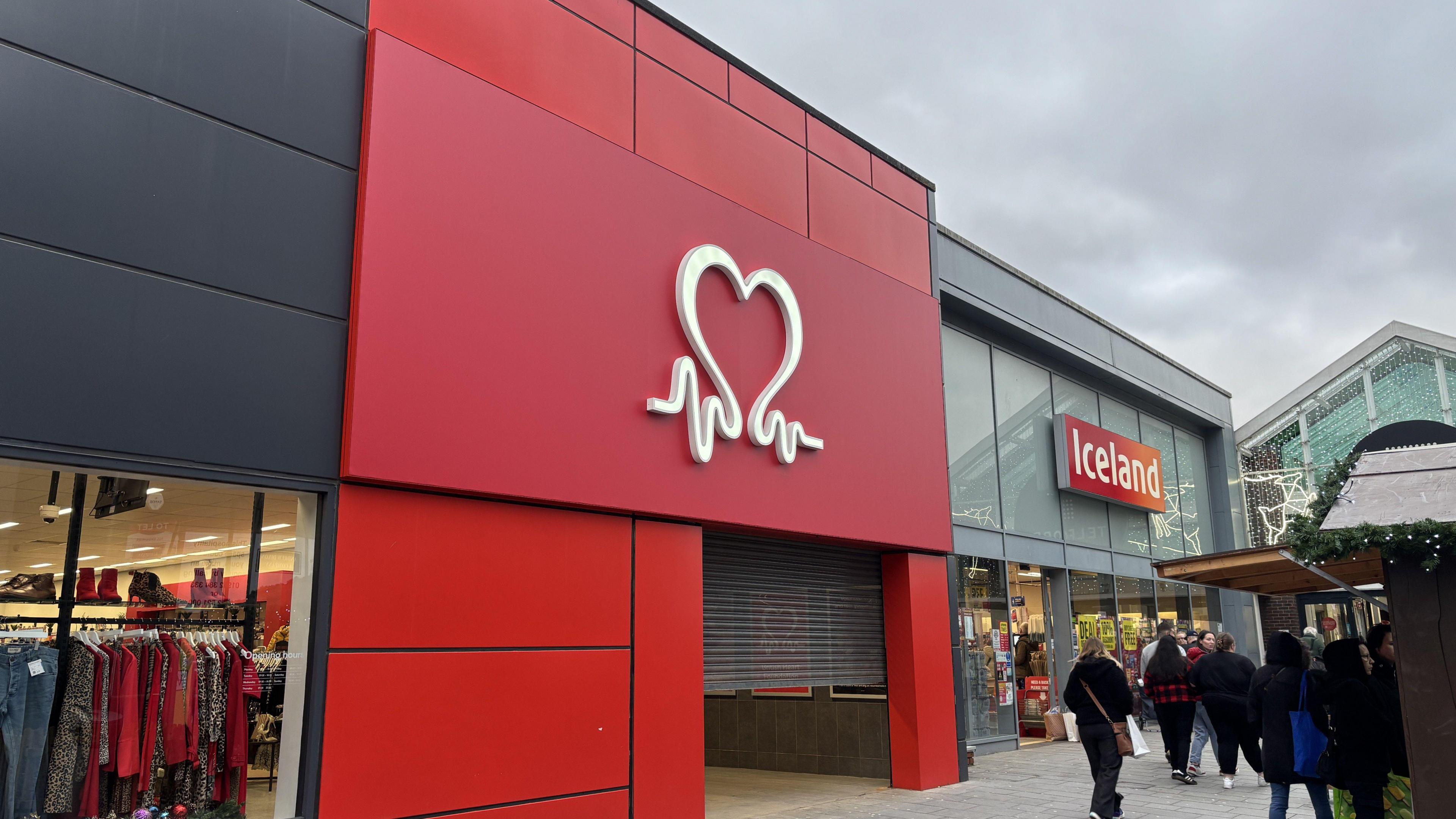 The front entrance of the new store. It is dark grey with large windows. Above the door, the panelling is red, and the white heart logo for the charity is over the door.