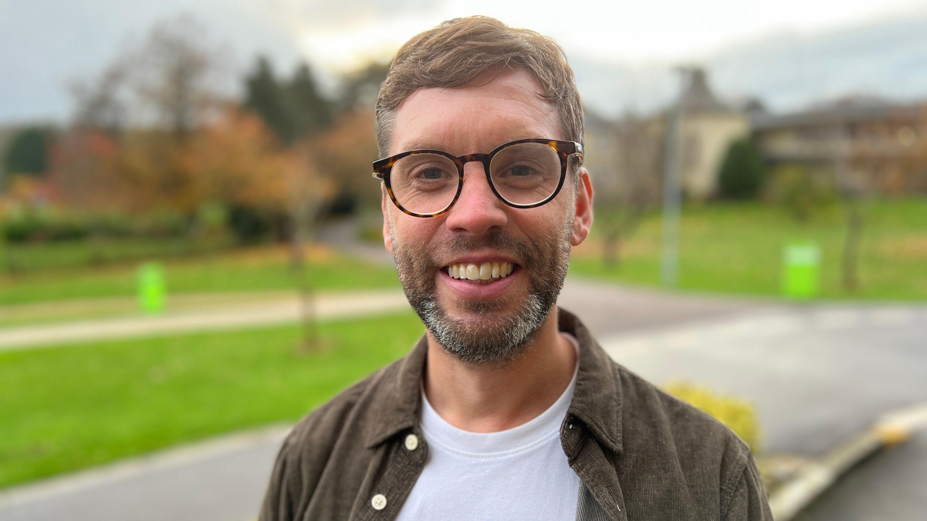 A person wearing a white T-shirt and an open brown shirt stands outdoors on a paved area with green grass, trees, and a building in the background. 