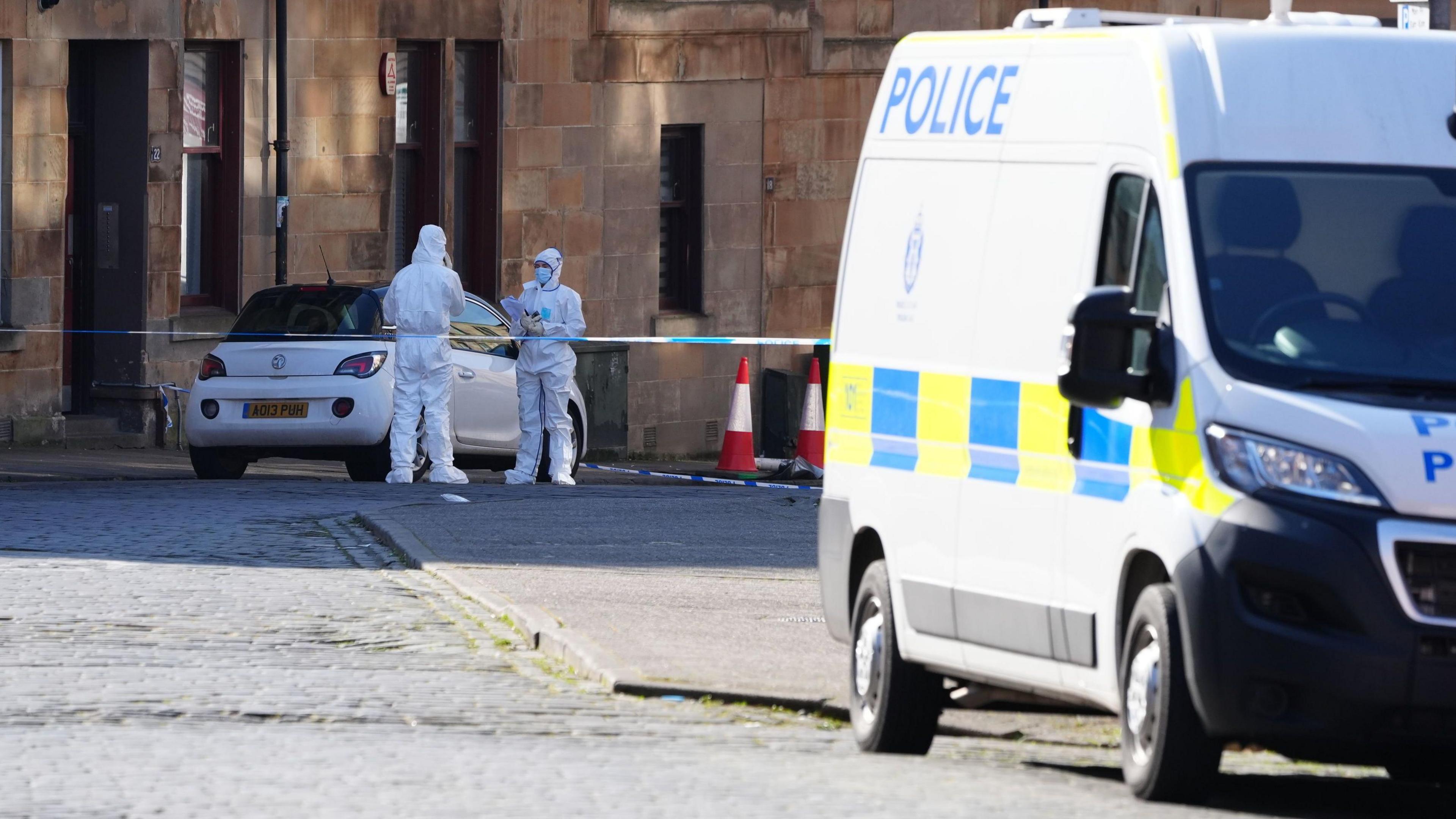 a white van with police Scotland logos on it, parked in front of crime scene tape. Two forensic officers in white suits stand in front of a white car.