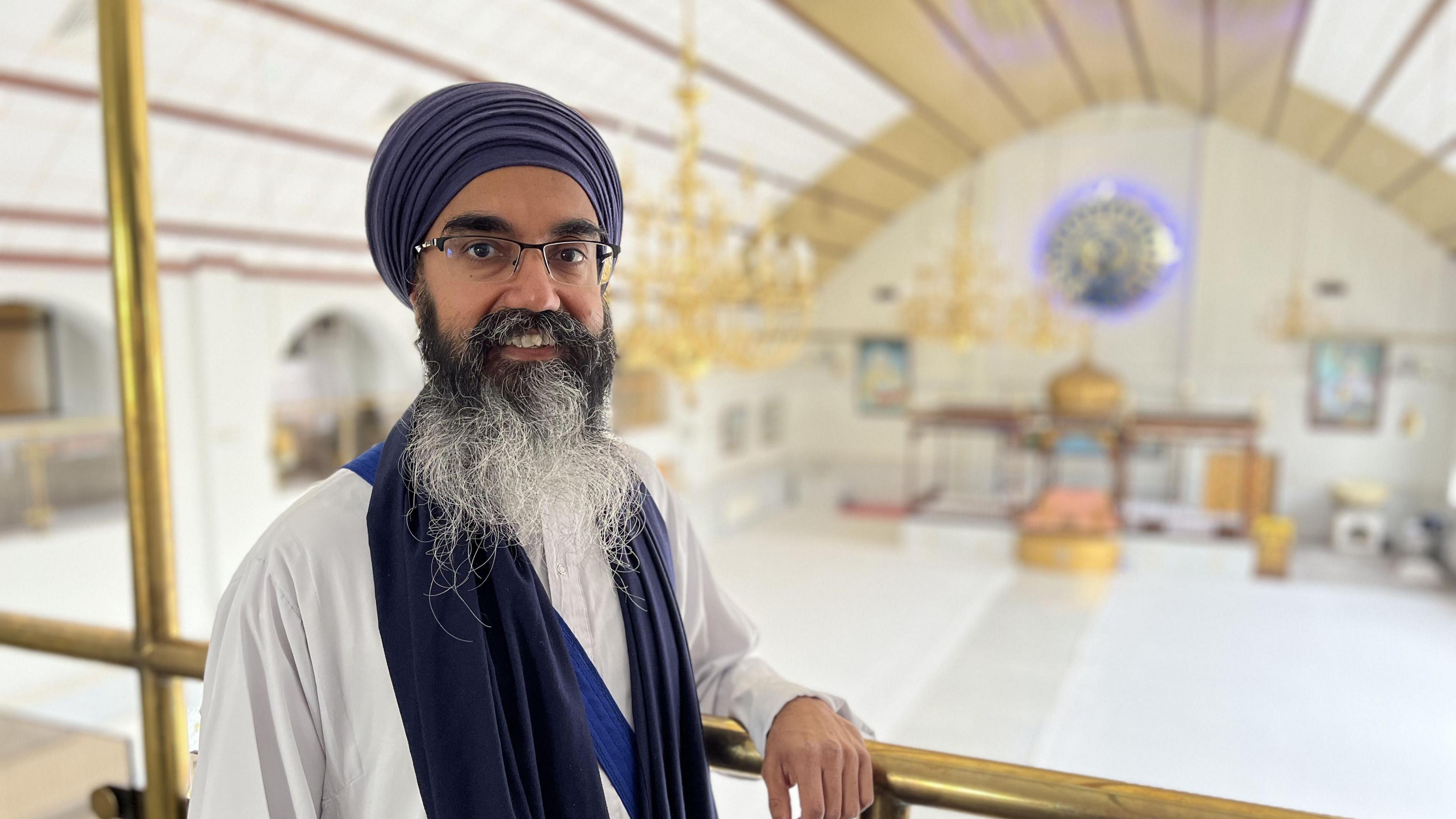 A man in a purple turban and white clothes with a beard and glasses smiles at the camera