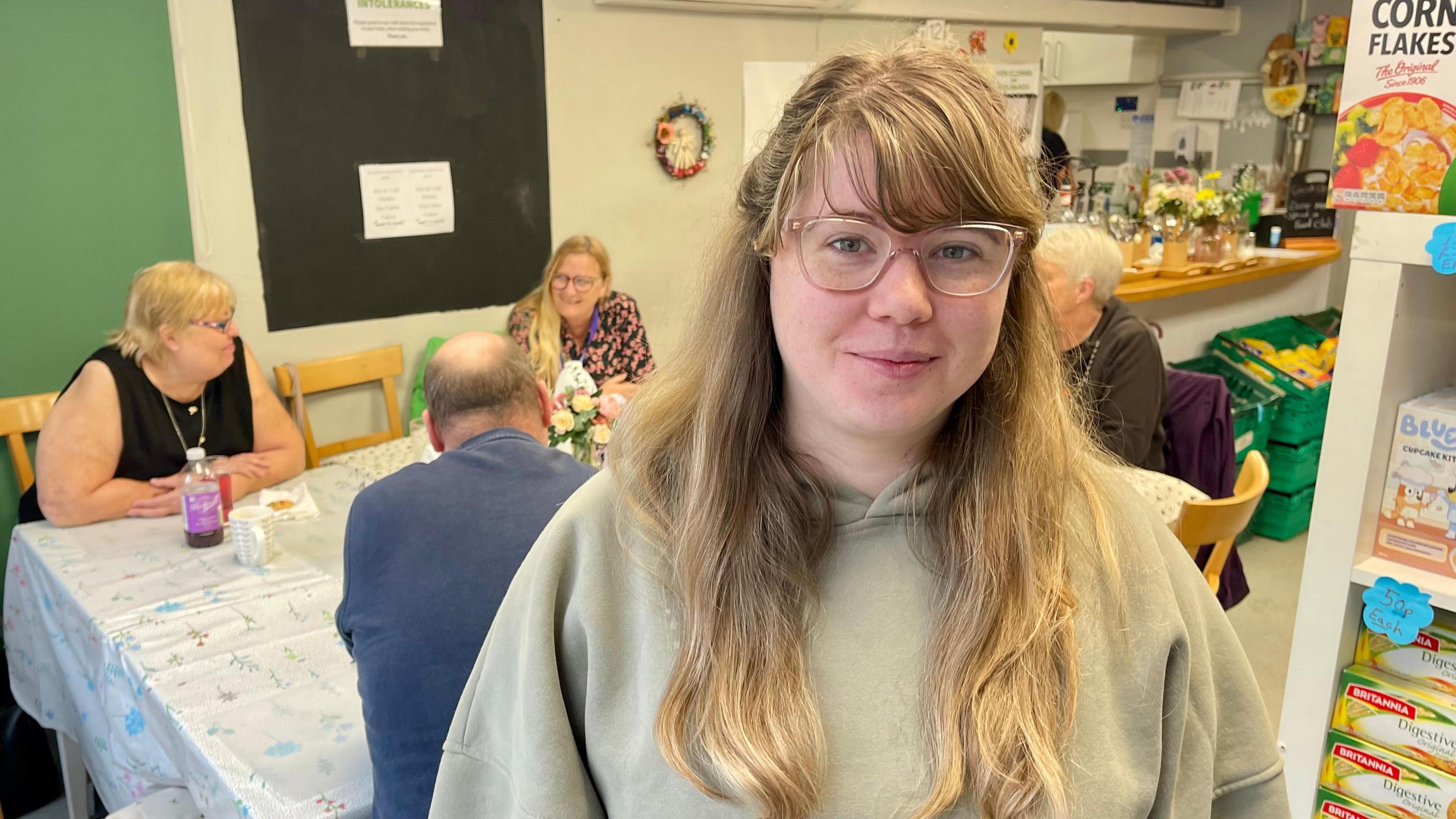 A woman with long fair hair, glasses and a light green hoodie is standing in a cafe. Behind her is a large table which three women and one man are sat around talking. A chalkboard is on the wall with a couple of laminated posters taped to it. A cafe counter can be seen in the corner of the room with green pallets in front of it. To the right of the woman are shelves packed with food products.