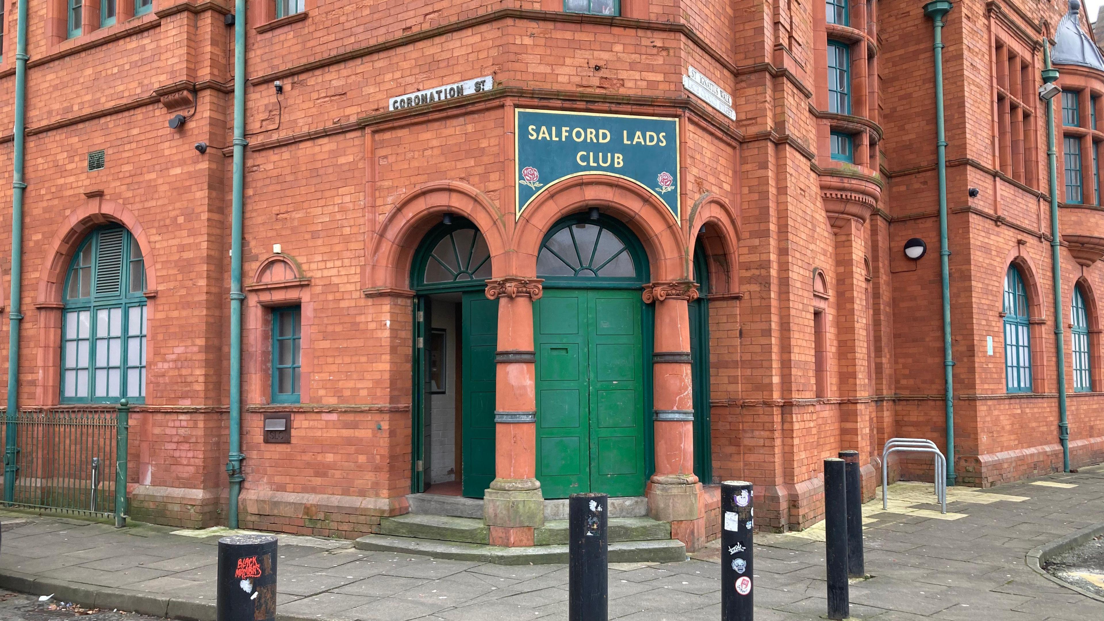 A red brick building with a green door and a sign that says Salford Lads Club in green over the door