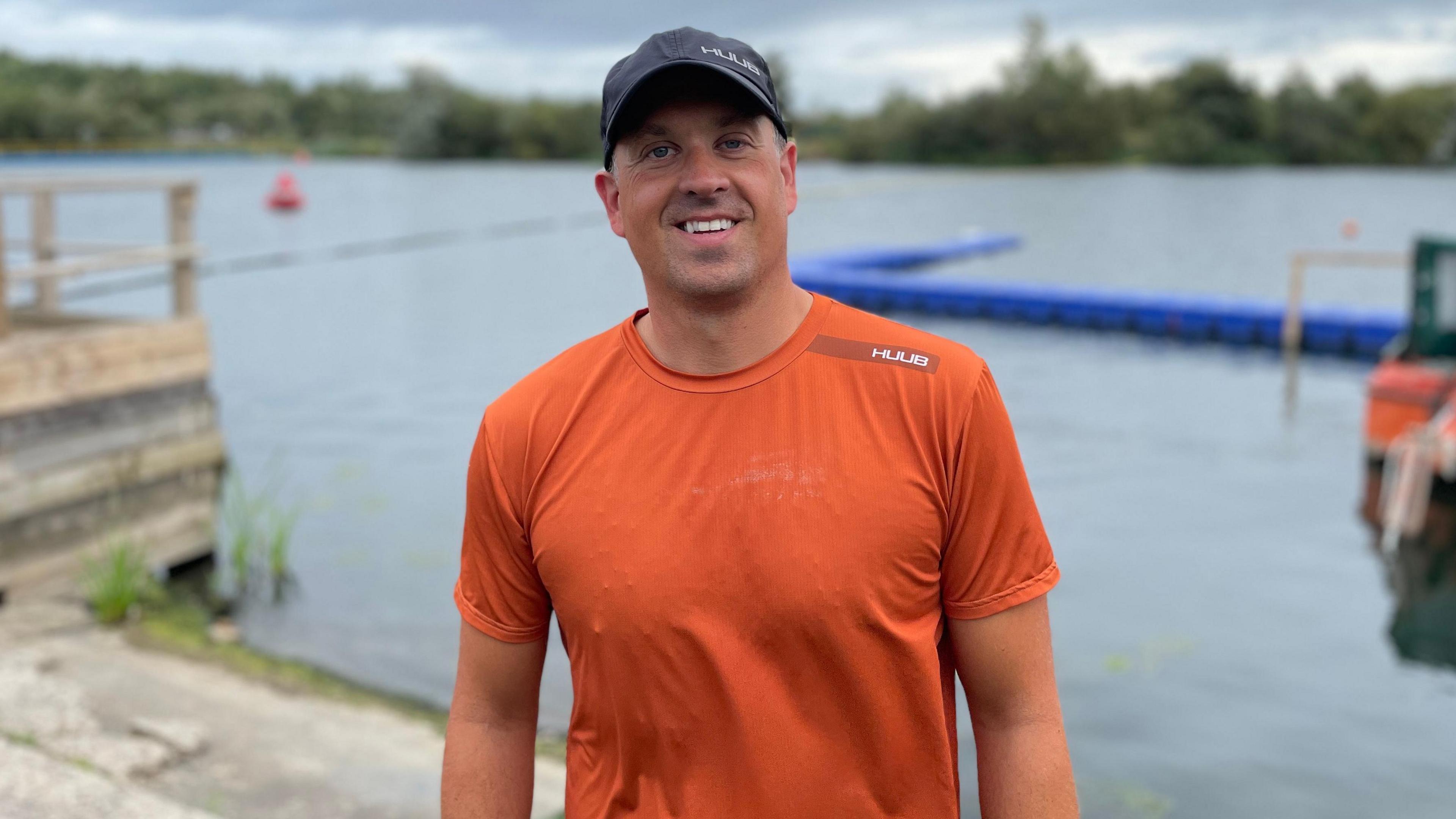 Man in dark blue baseball cap and orange t-shirt in front of a lake