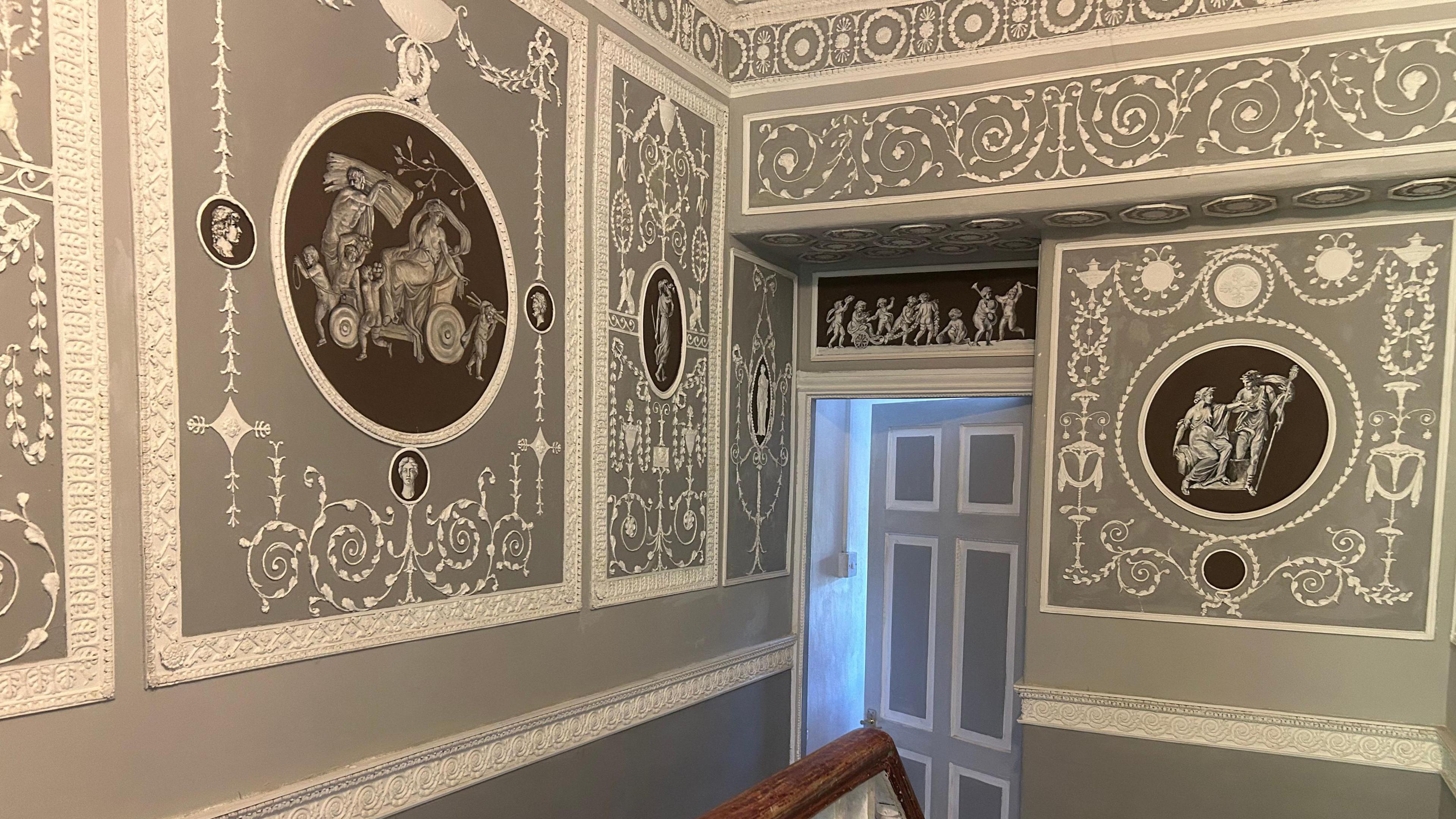 Close-up image of the upstairs wall in the house. They are painted grey with intricate white neoclassical detailing in rectangular panels, featuring a variety of creative designs in black circles, including 18th Century portraits of Roman looking figures. 