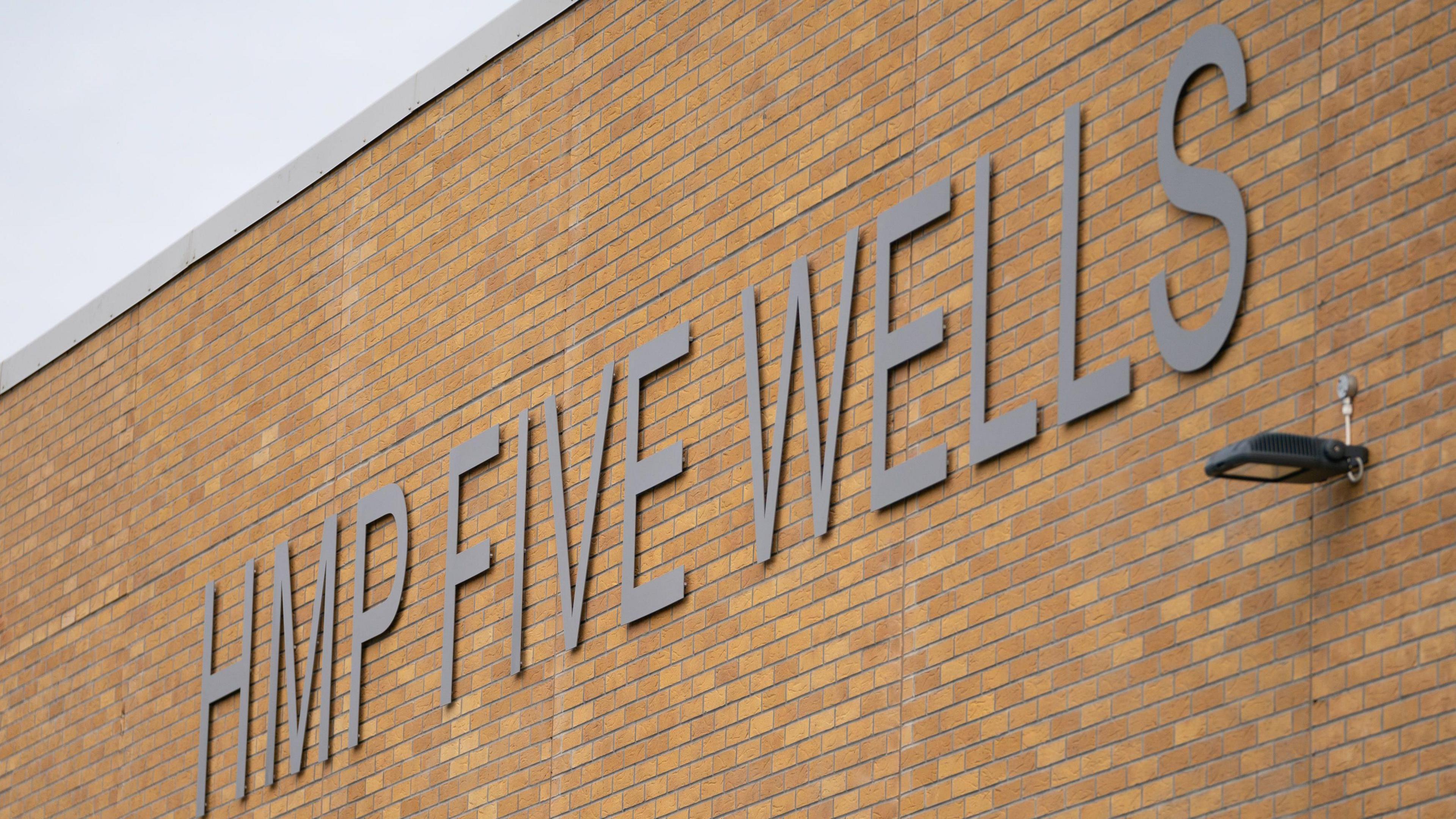 A sign for HMP Five Wells displayed on a large brick wall.