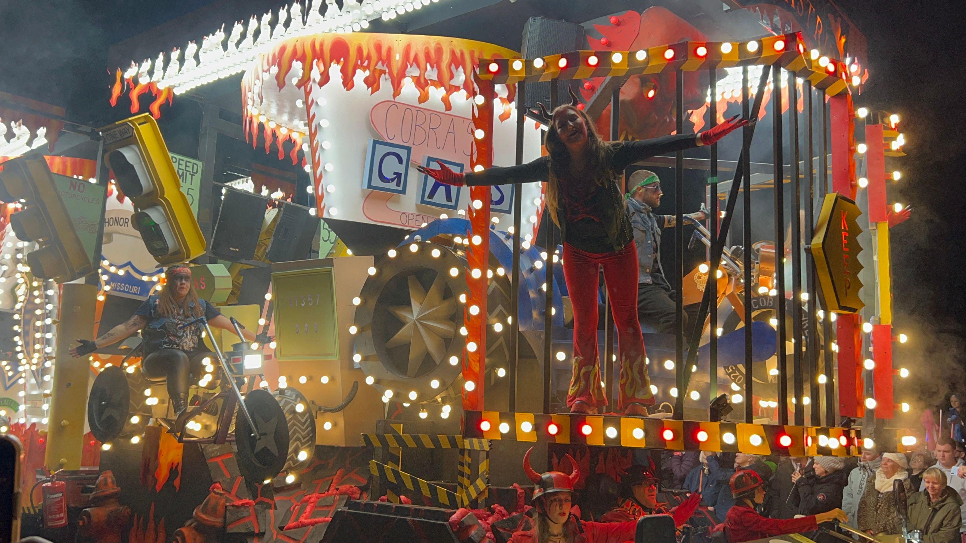 An illuminated carnival cart. There is a woman stood at the front with her arms stretched out.
