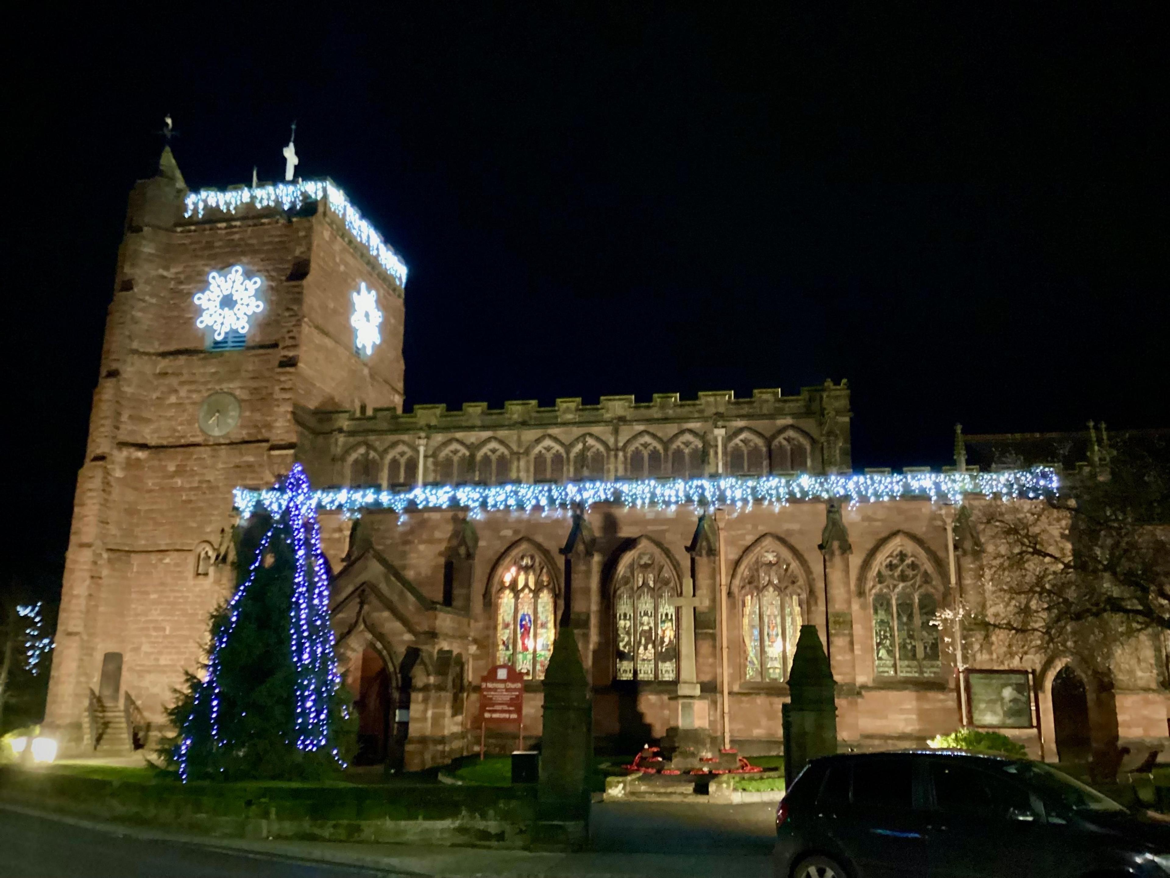 A church at night with an illuminated tree in the foreground, and lights along the roof edge and around  the tower.