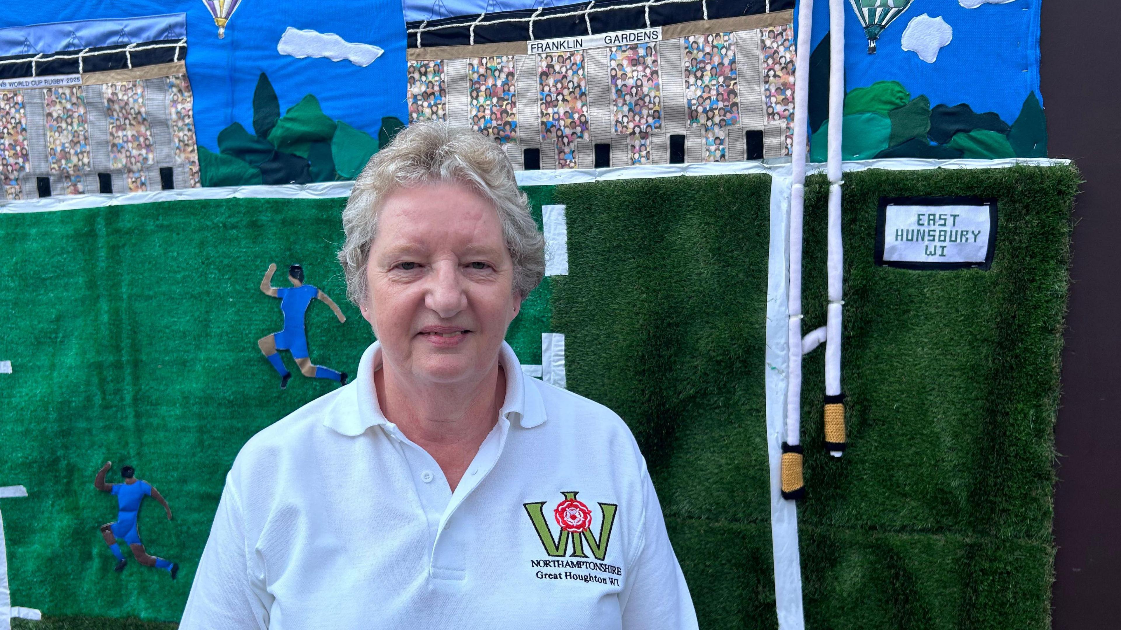 A woman in a white polo shirt stands in front of a knitted green mural with blue knitted rugby players on it.
