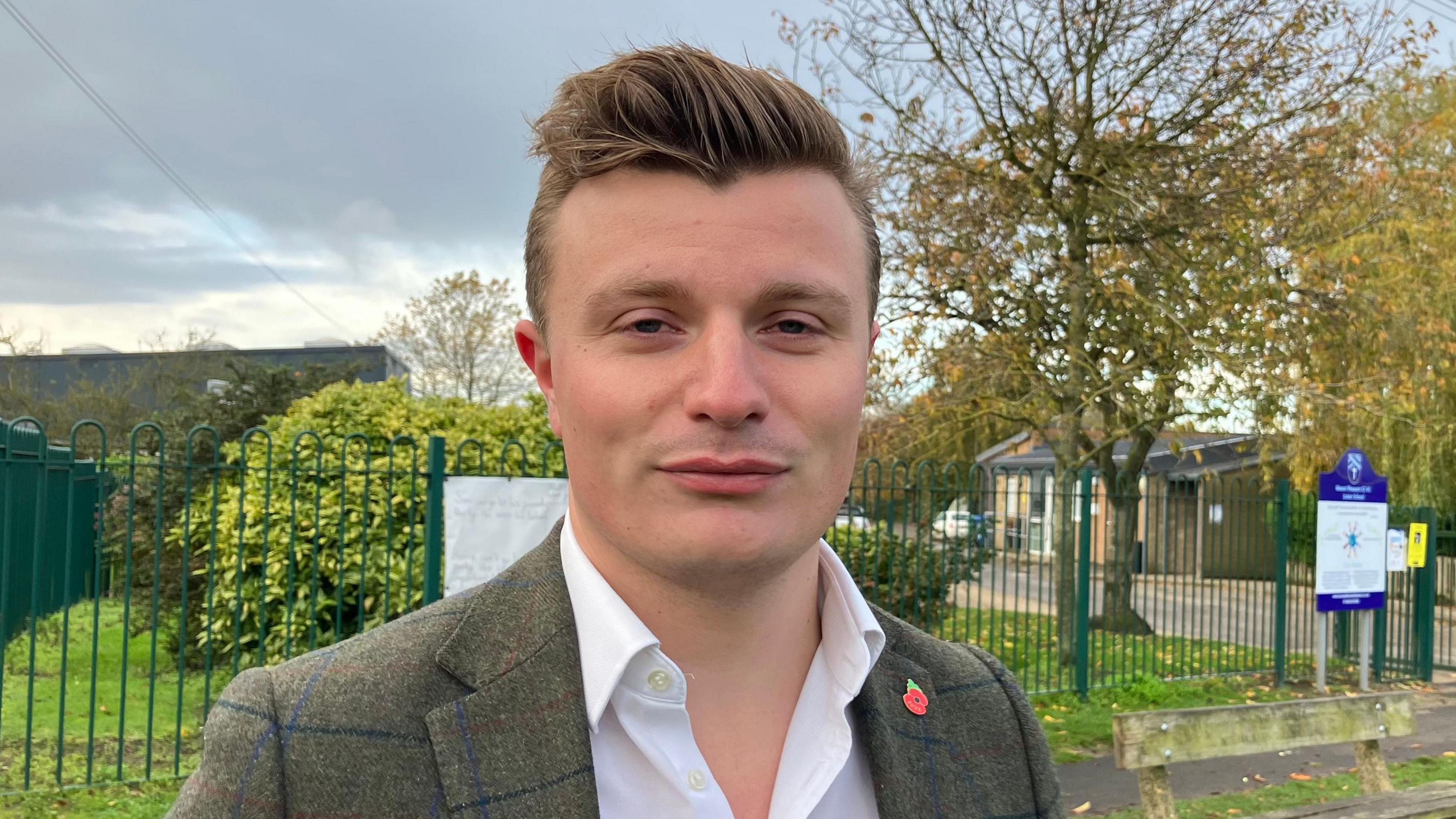 A young man with fair hair, wearing a white shirt and tweed jacket.