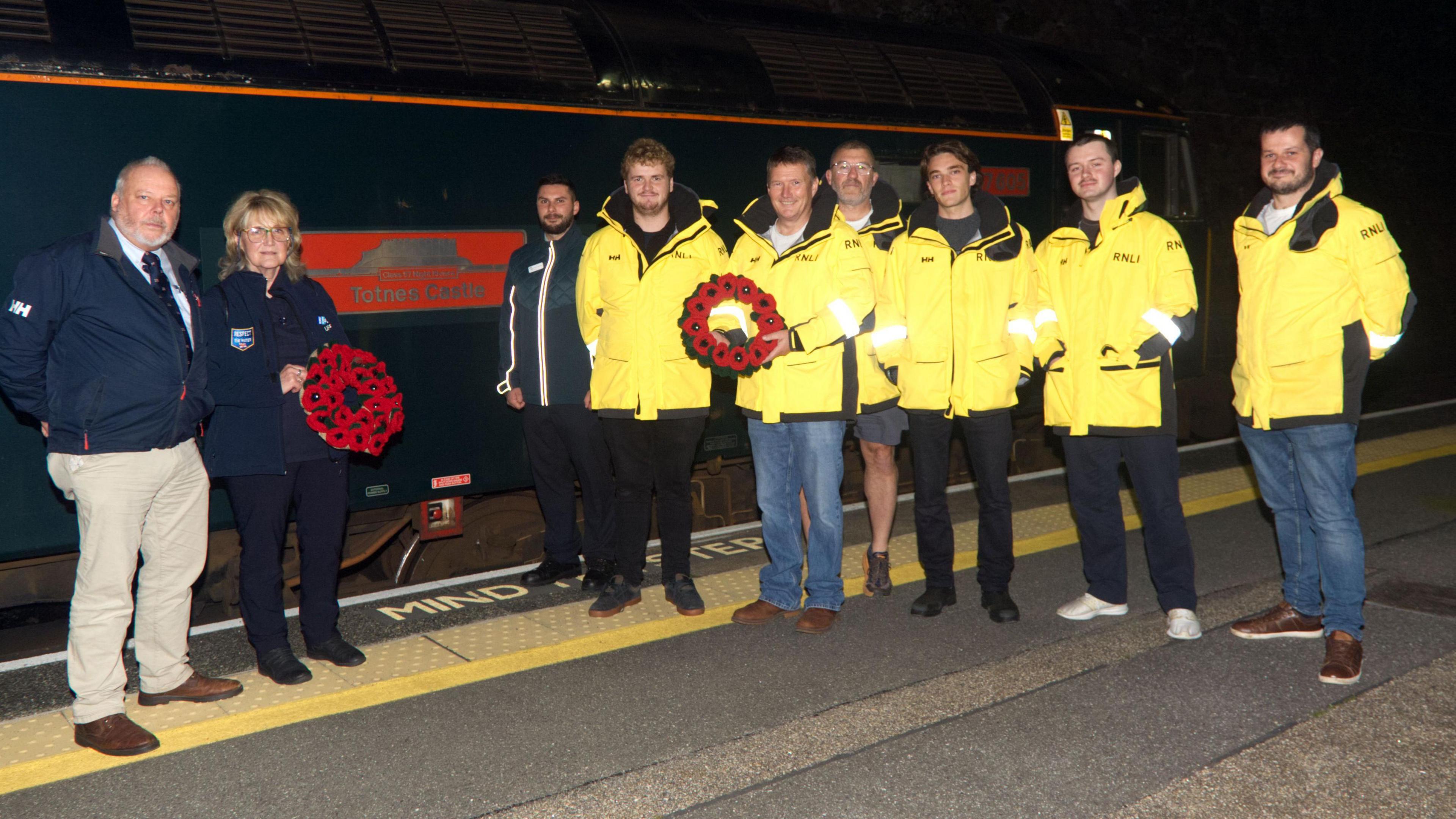 Six people in yellow coats and three in darker jackets. A woman and a man are each holding a poppy wreath. They are standing on a platform next to the Totnes Castle sleeper train.