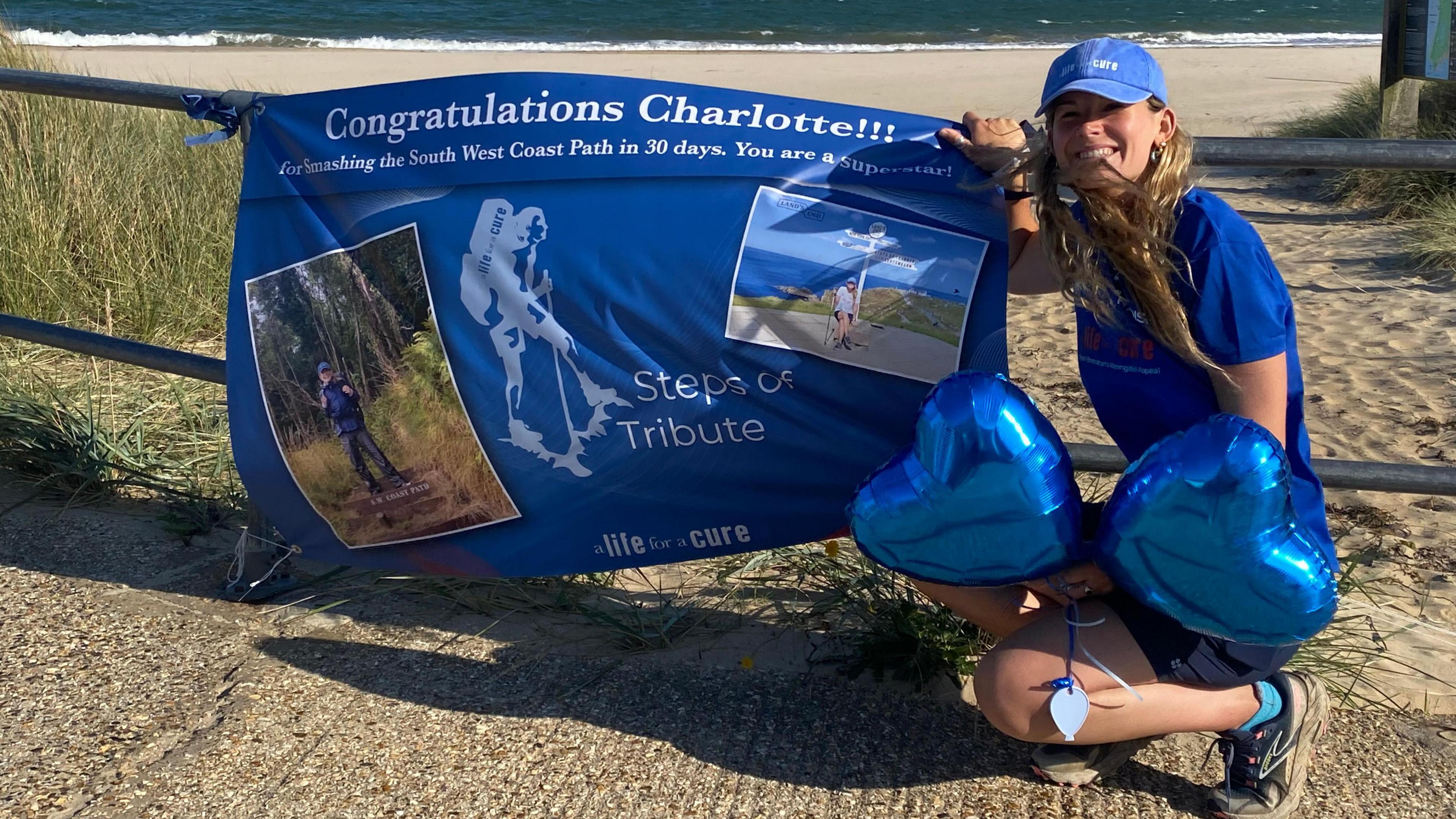 Charlotte, wearing blue, and holding two blue heart balloons, crouched next to a banner reading "congratulations Charlotte!!!"